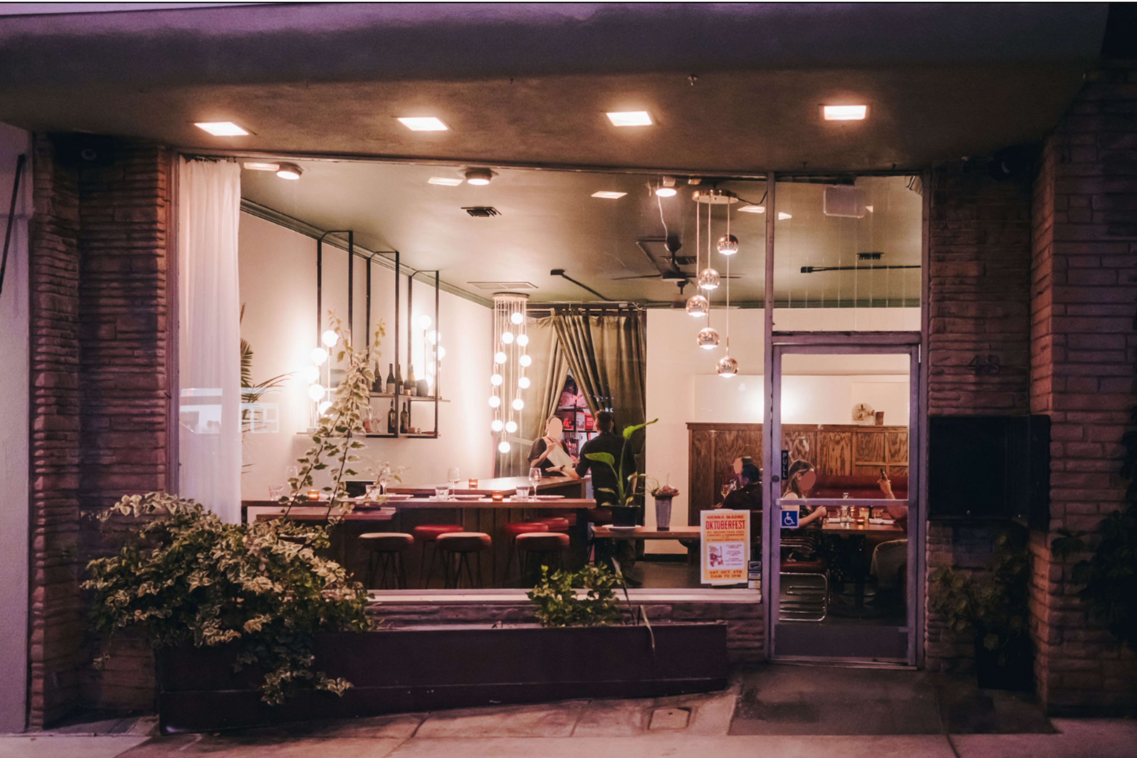 The image shows a modern restaurant exterior with a large glass window, featuring indoor seating and decorative lighting.