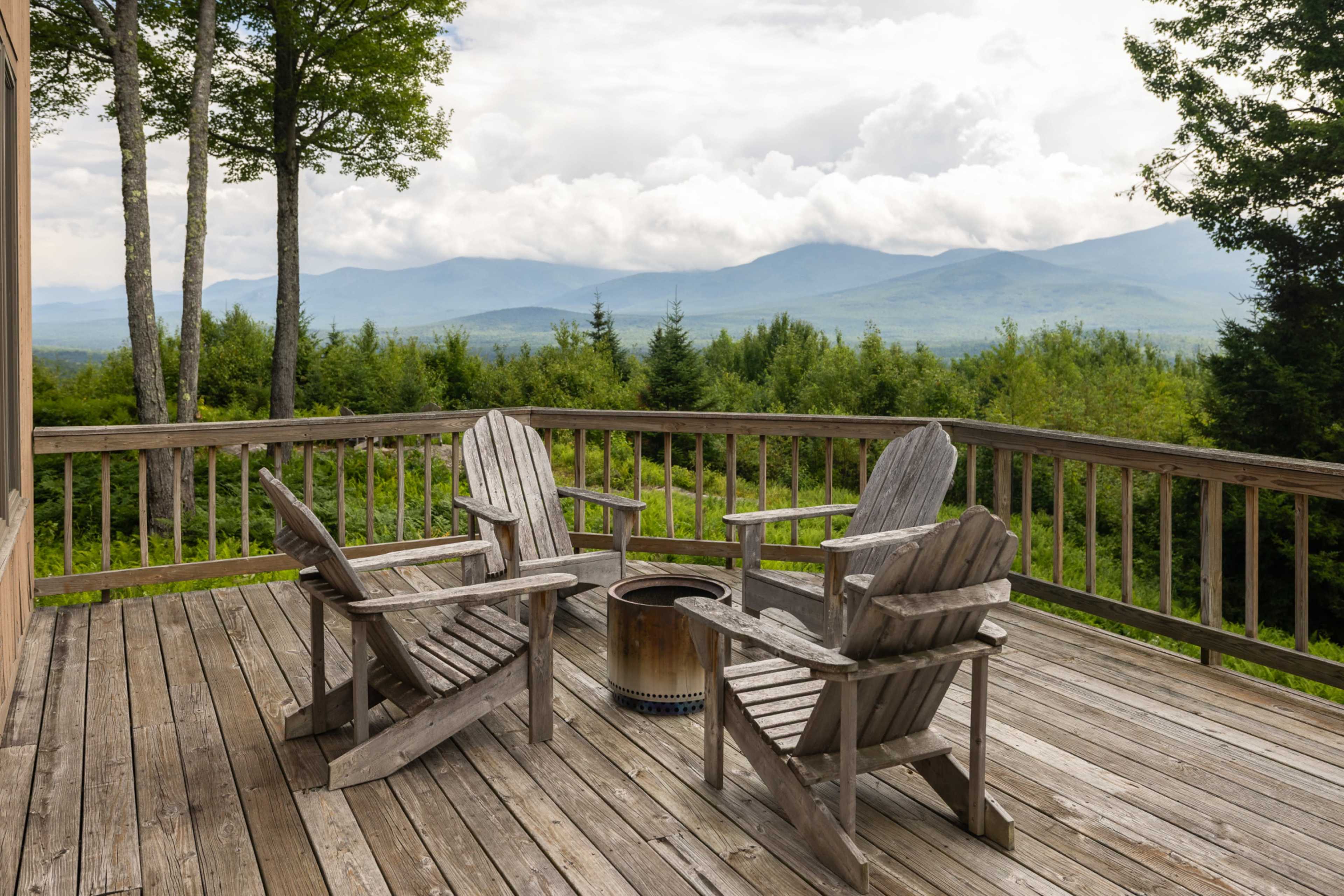 The image shows a wooden deck with four Adirondack chairs arranged around a metal fire pit, overlooking a mountainous landscape under a cloudy sky.