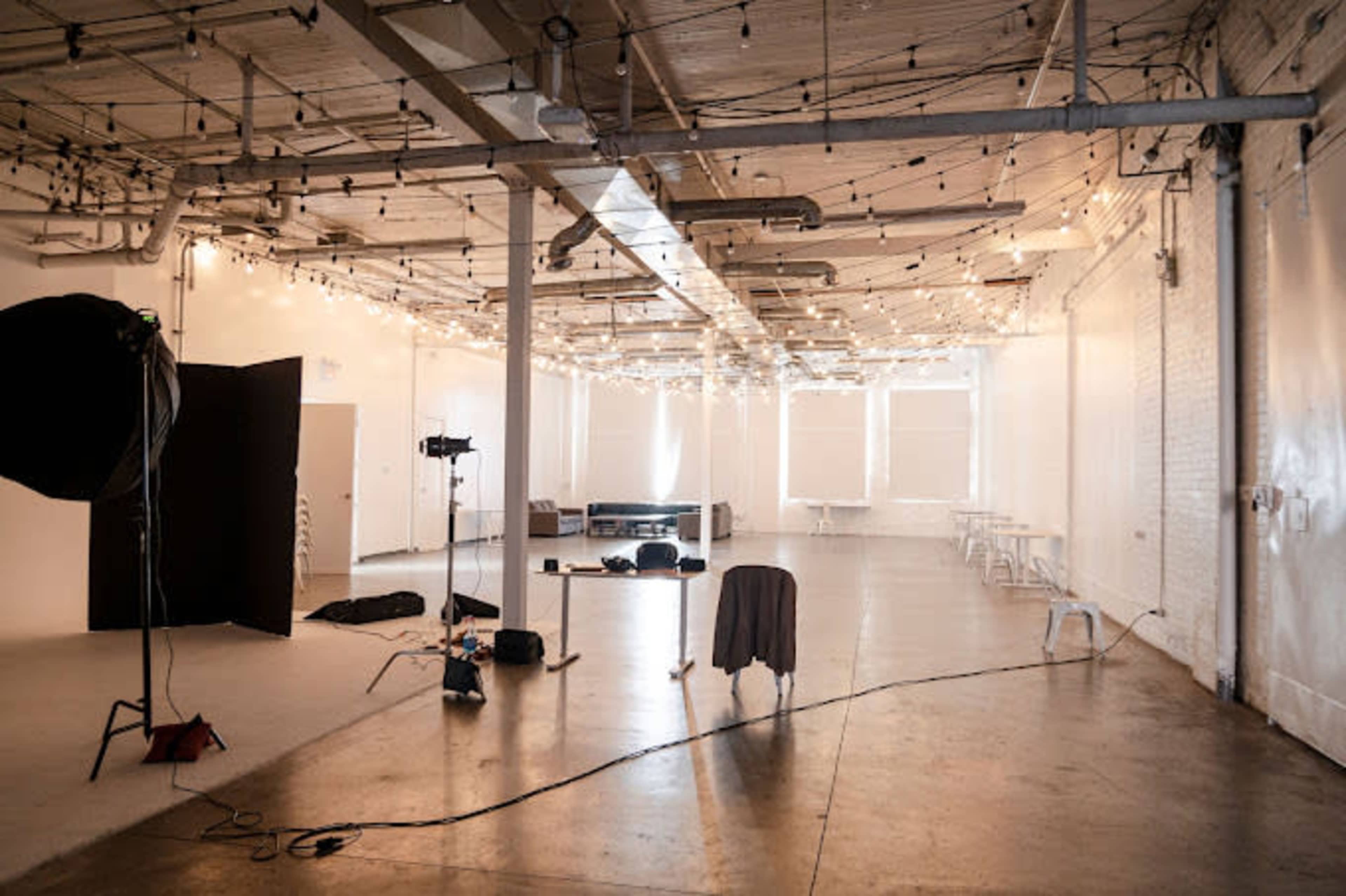 A spacious, well-lit studio with string lights overhead, featuring a large central area, a few pieces of furniture, and various studio equipment.