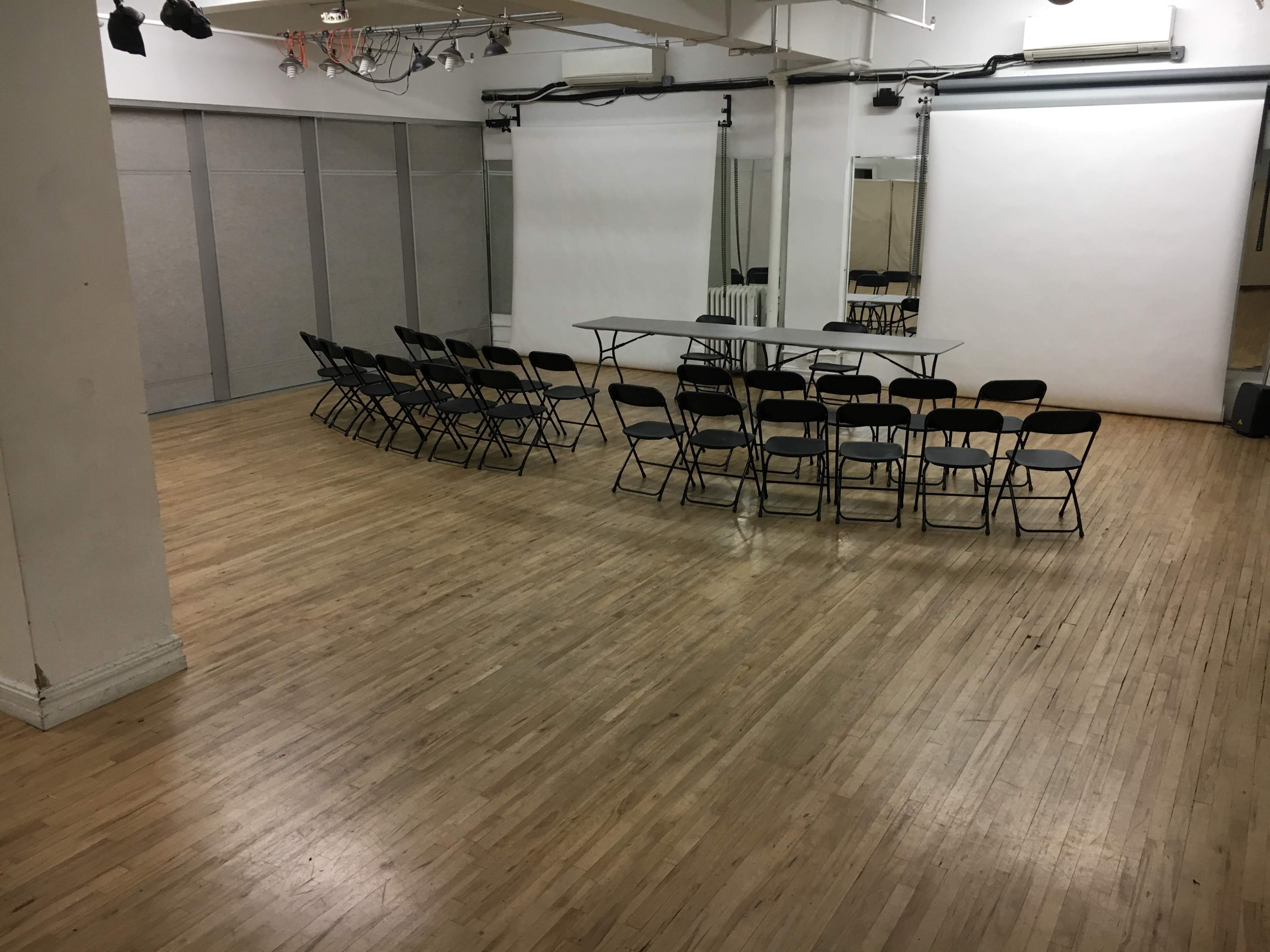 A spacious room with a wooden floor, featuring a row of black chairs arranged around a central table, along with two large white screens at the front.