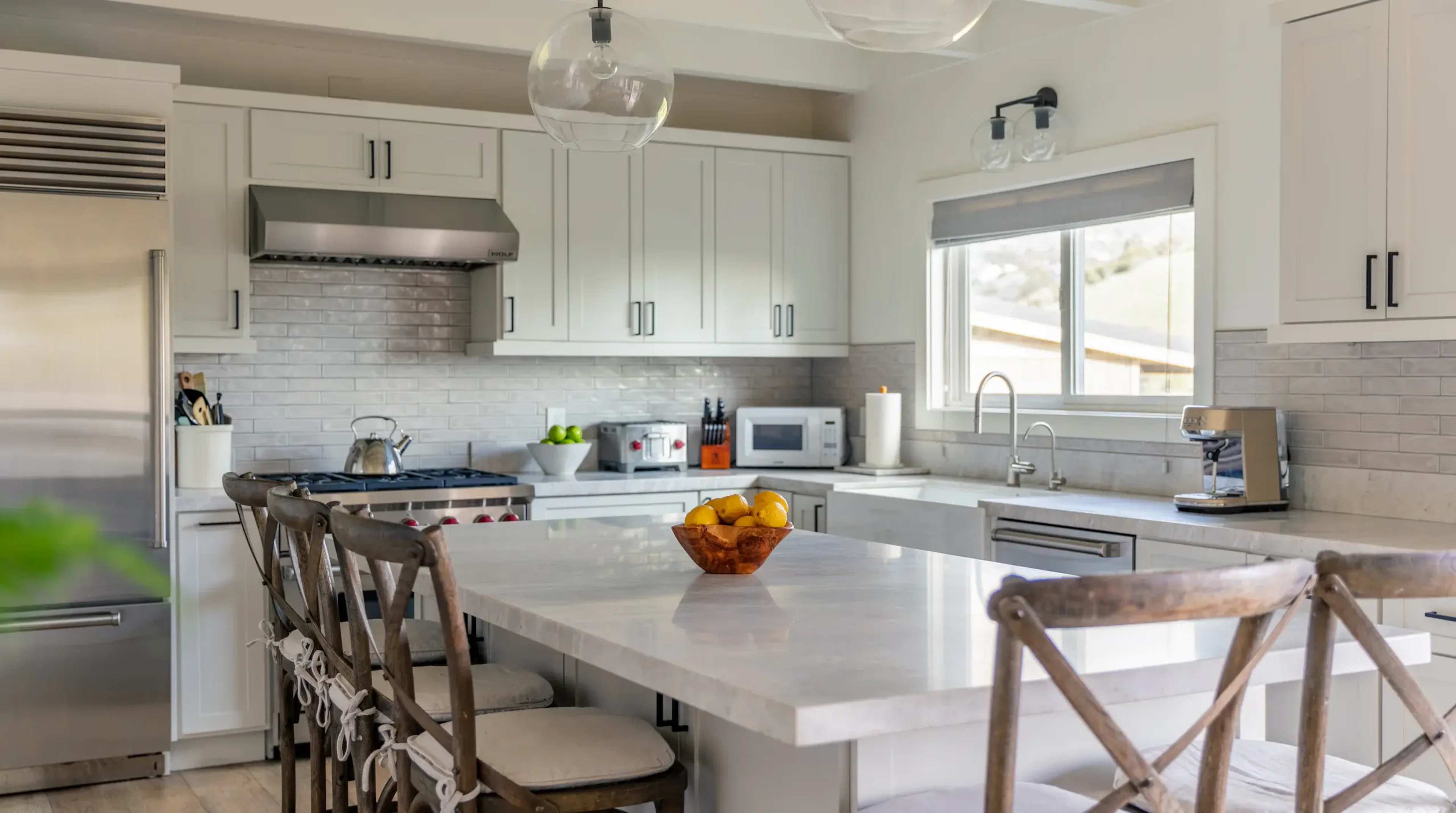 A modern kitchen features white cabinetry, a central island with a marble top, and a bowl of fruit on the table.