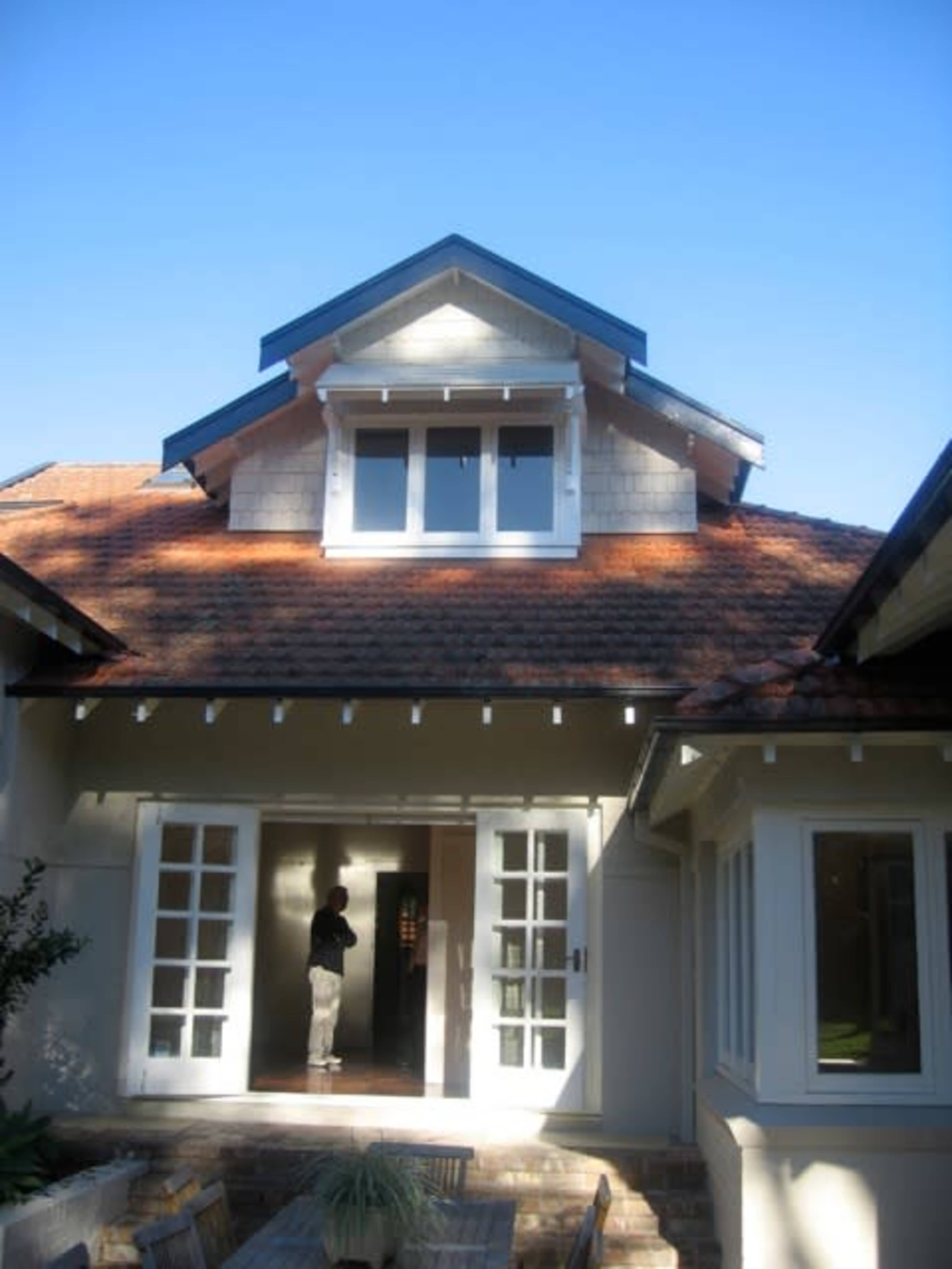 A person stands in the doorway of a house with a gabled roof and multiple windows under a clear blue sky.