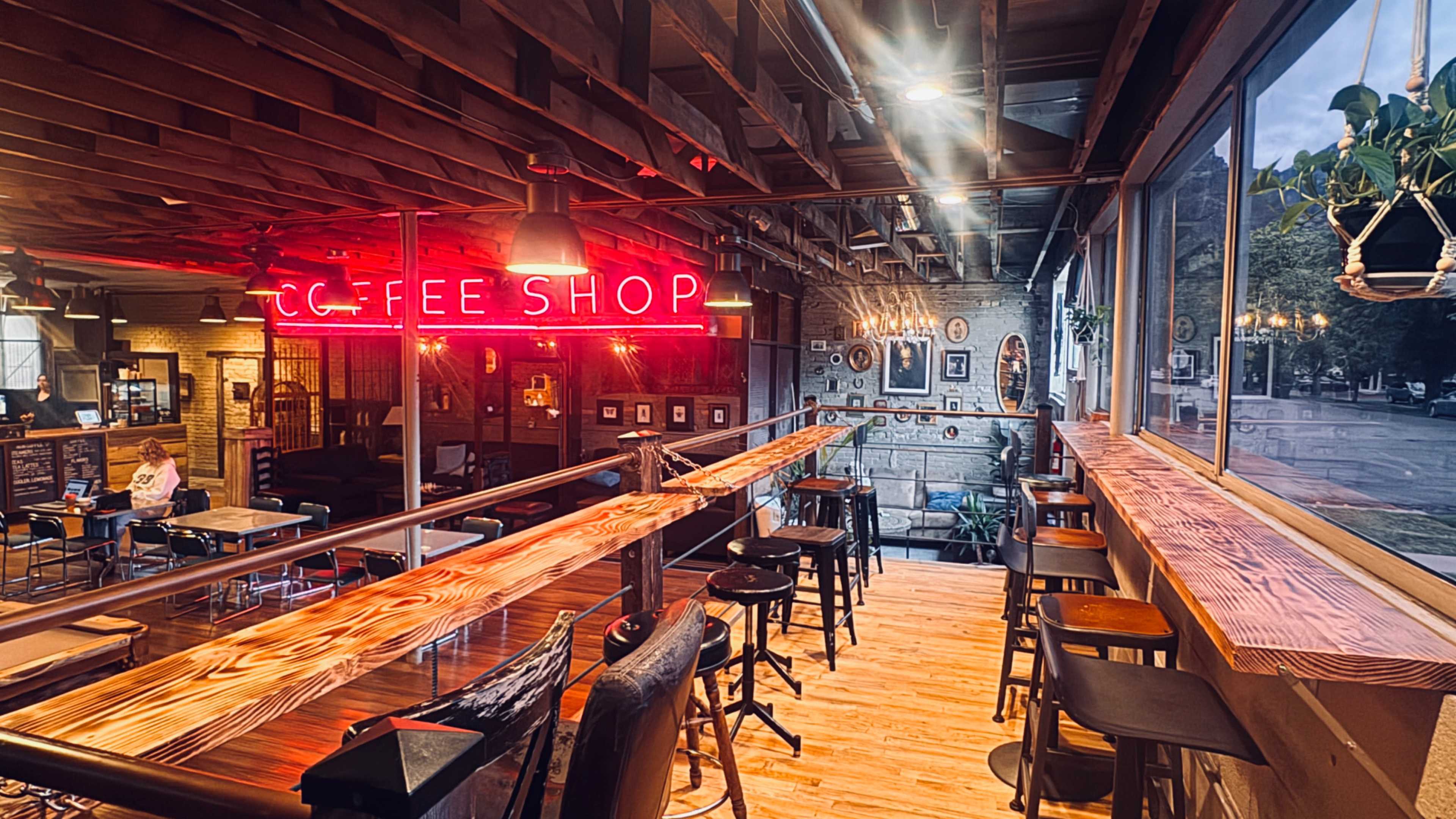 The image shows the interior of a coffee shop with a long wooden counter, bar stools, and neon signage that reads "COFFEE SHOP" against a backdrop of exposed brick walls and large windows.