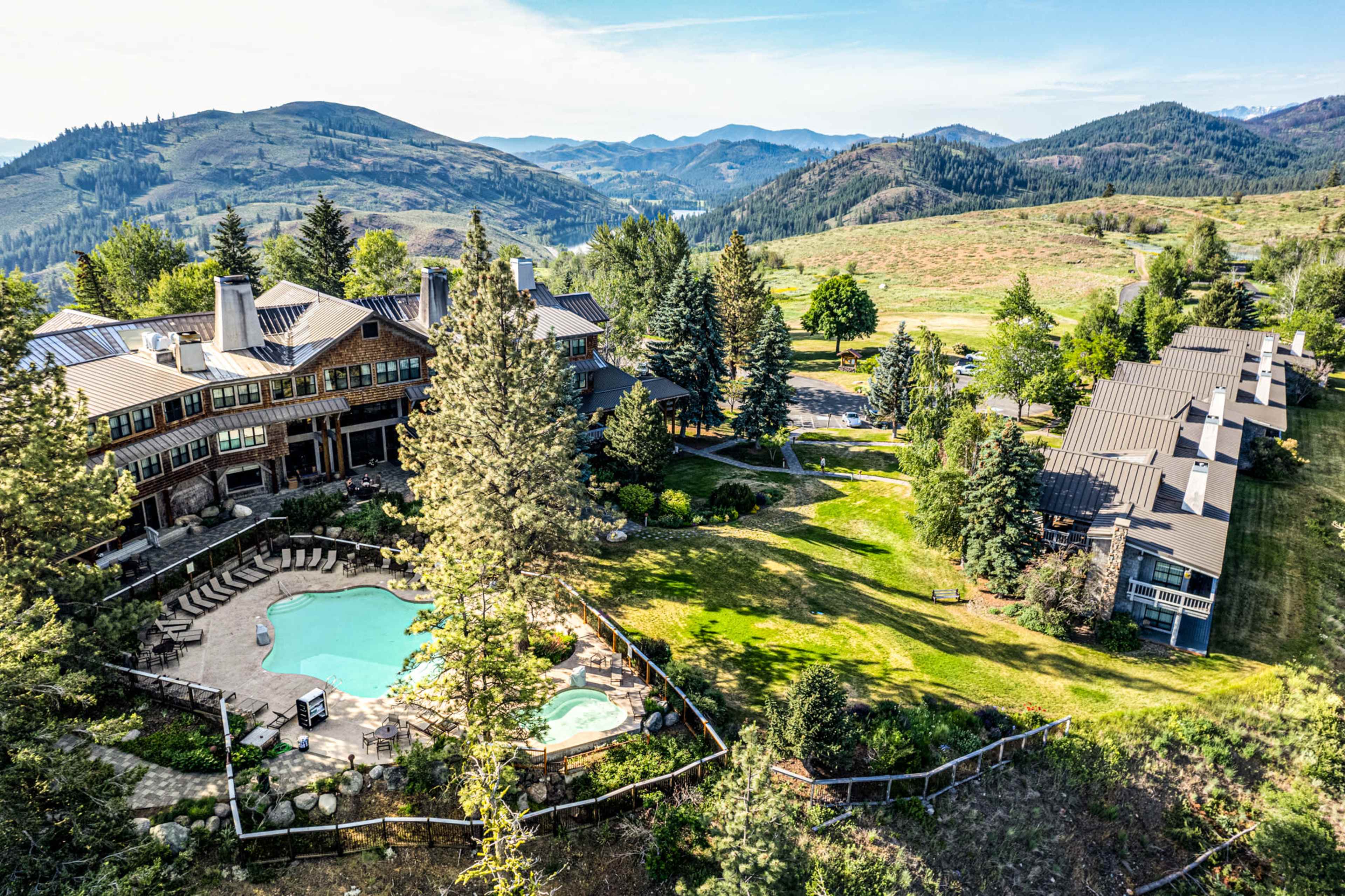 Aerial view of a large lodge with a swimming pool surrounded by trees and mountains in the background.