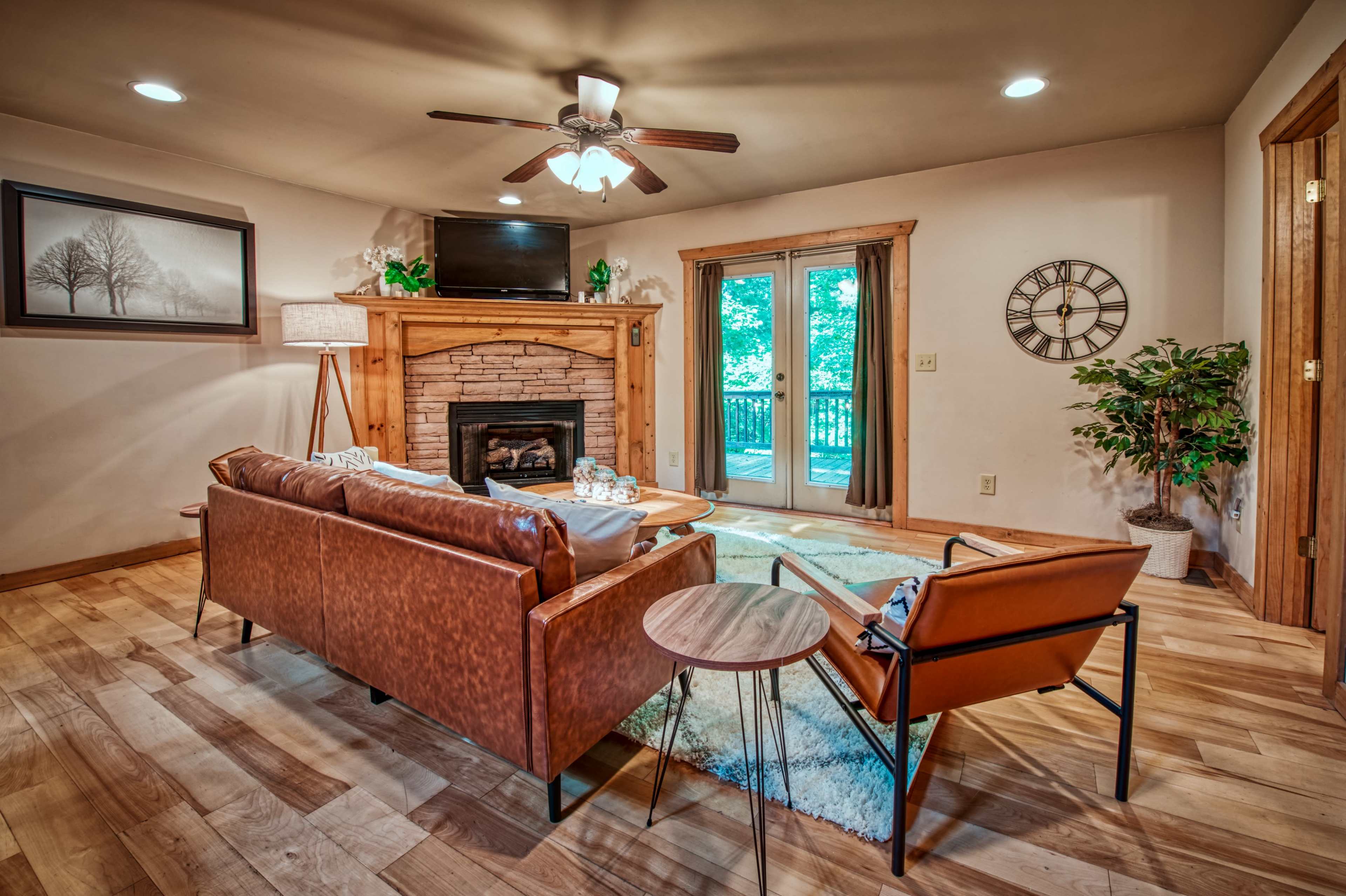 The living room features a leather sofa, a wooden coffee table, and a stone fireplace, with large windows opening to an outdoor view.