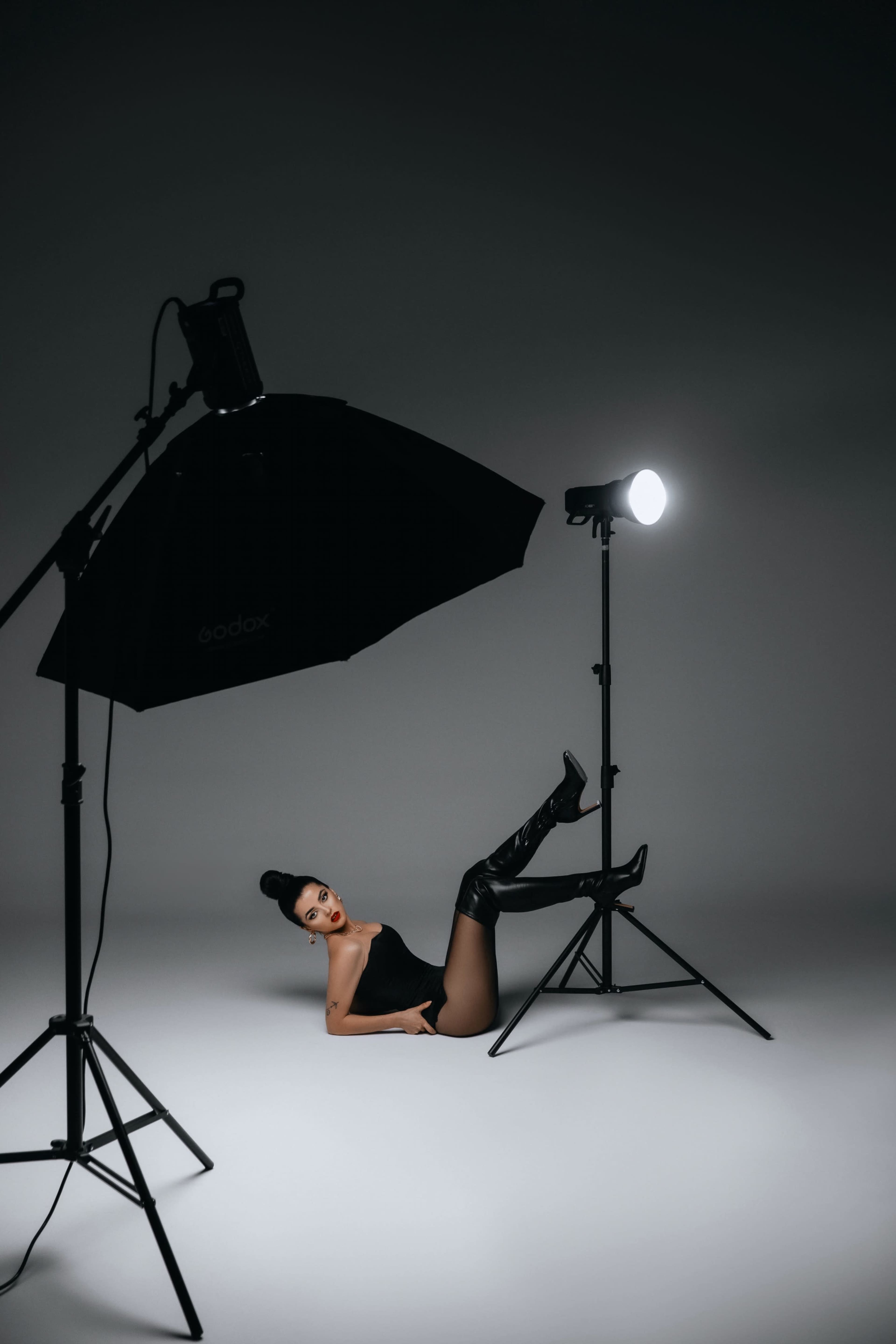 A model poses on a light-colored floor with one leg raised, while studio lighting equipment is positioned around her.