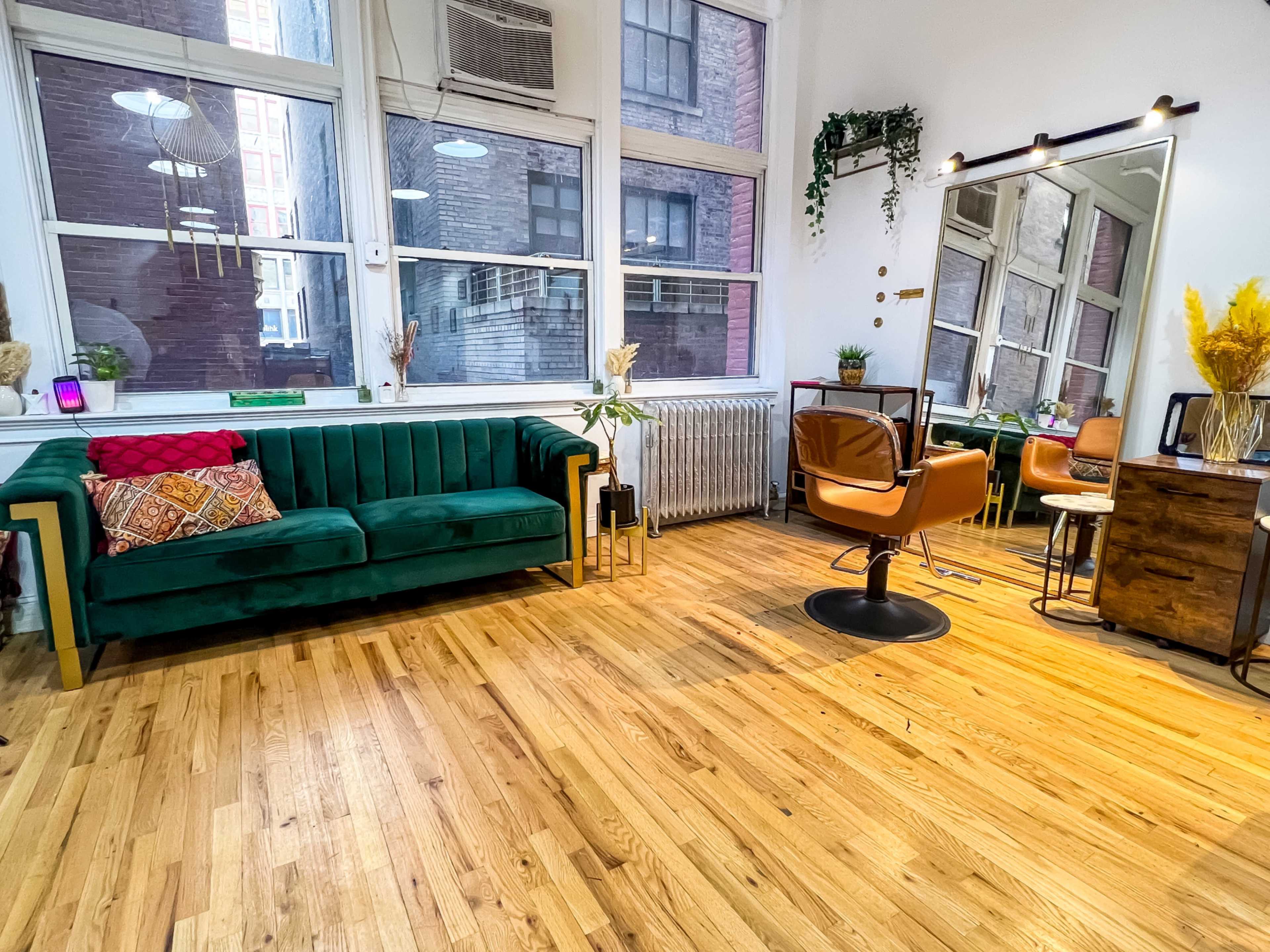 A stylish salon interior featuring a green couch, large windows, a mirror, and salon chairs with wooden flooring.