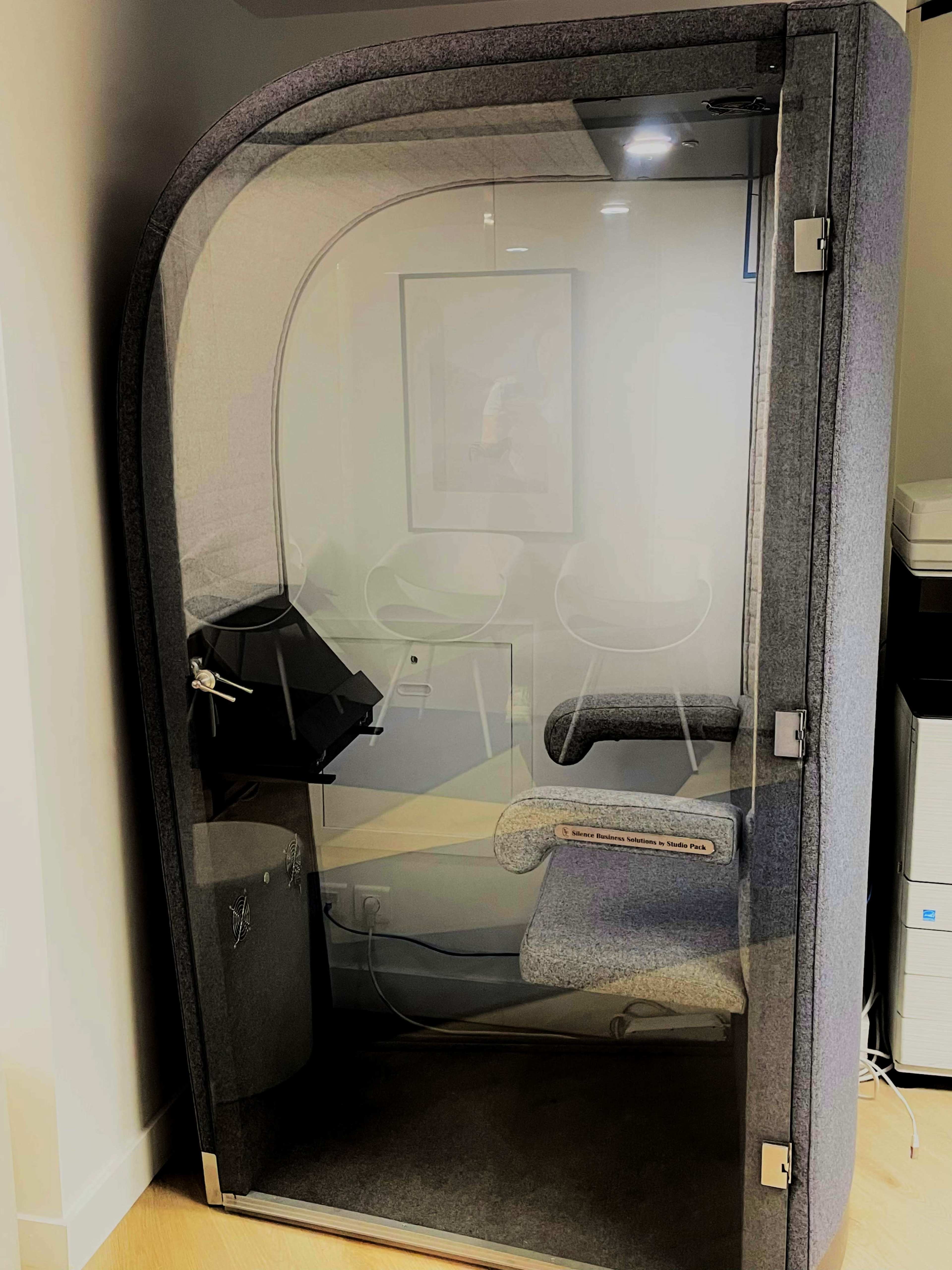 The image shows a soundproof phone booth with a comfortable chair and a small desk inside, enclosed in a modern design.