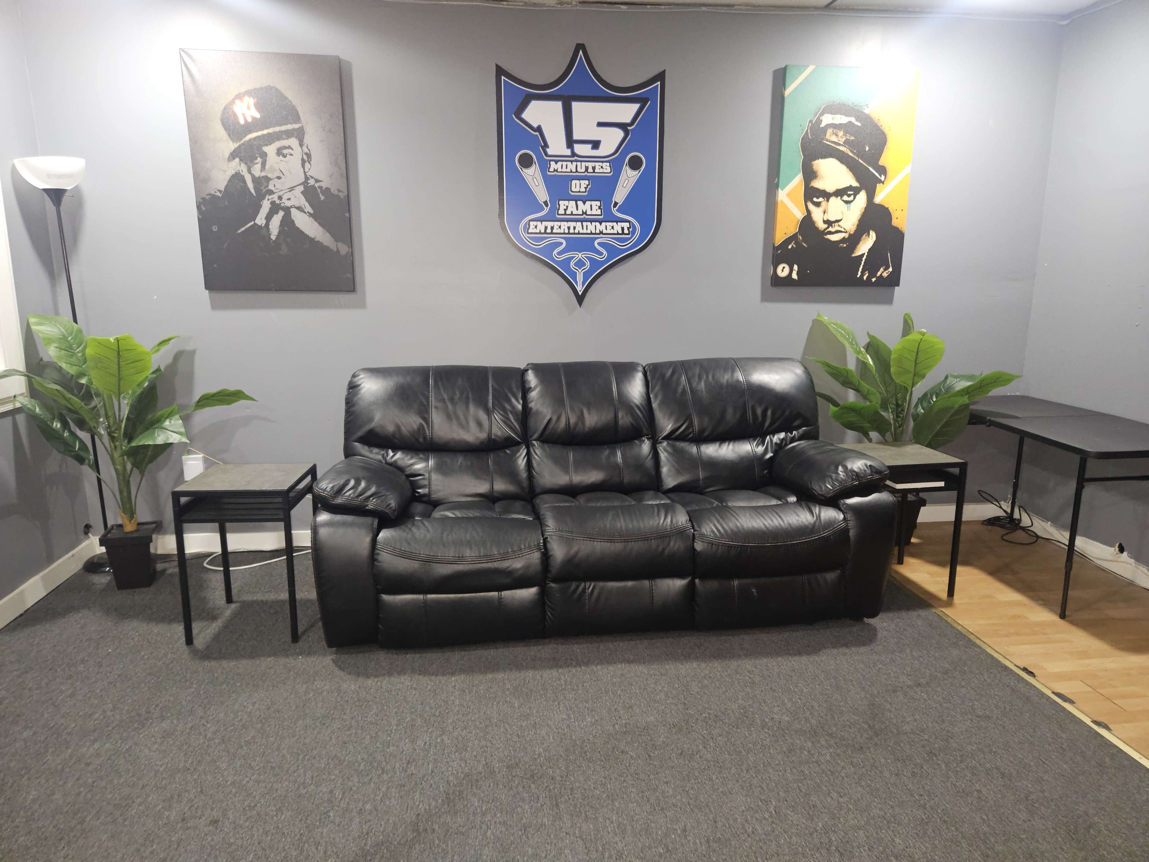 The image shows a modern room with a black leather couch, two small side tables, and two framed portraits on the walls, along with potted plants.