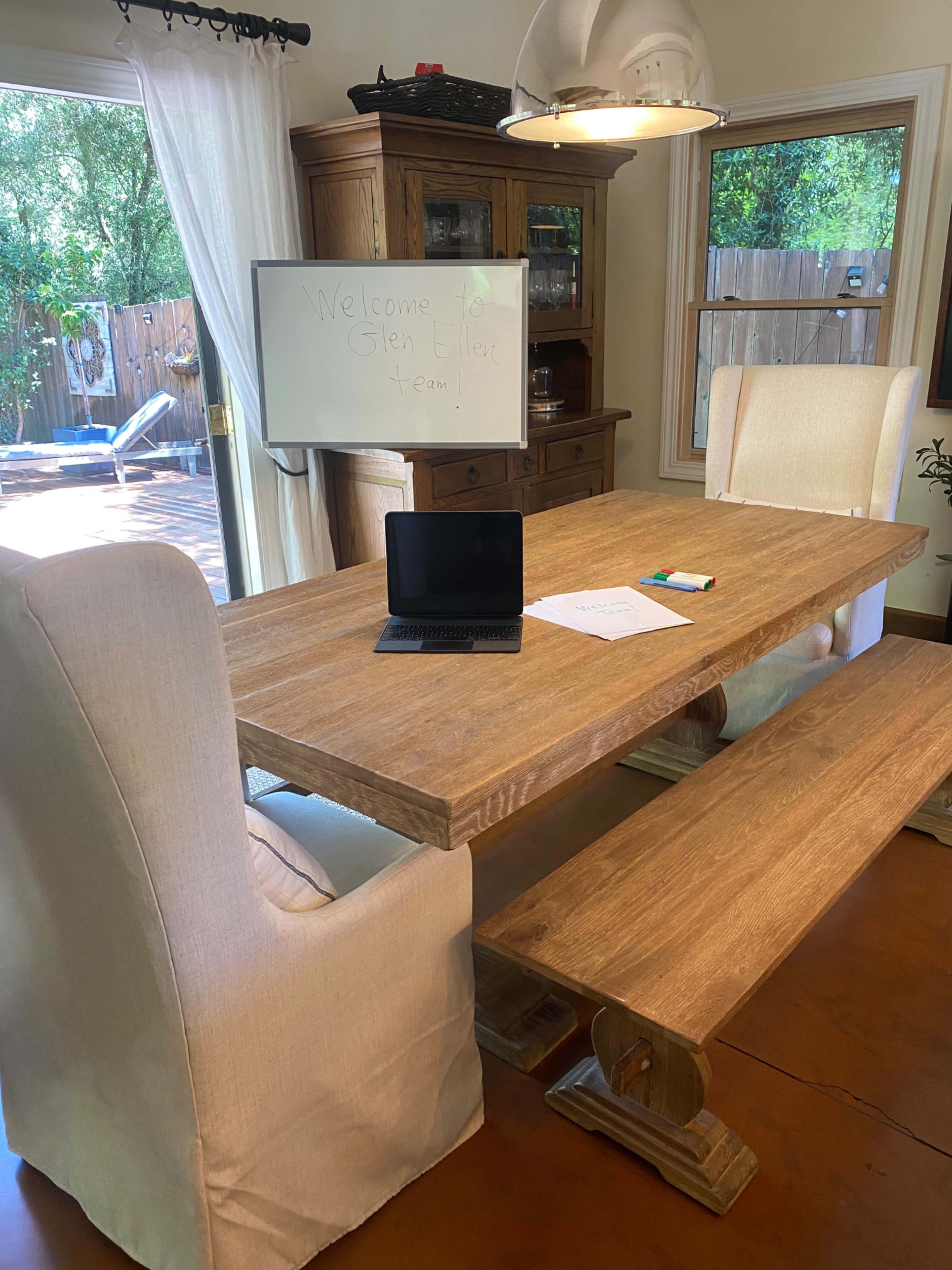 A wooden dining table is set with a laptop, papers, and colorful markers, surrounded by two upholstered chairs in a bright room with a chalkboard and a view of an outdoor patio.
