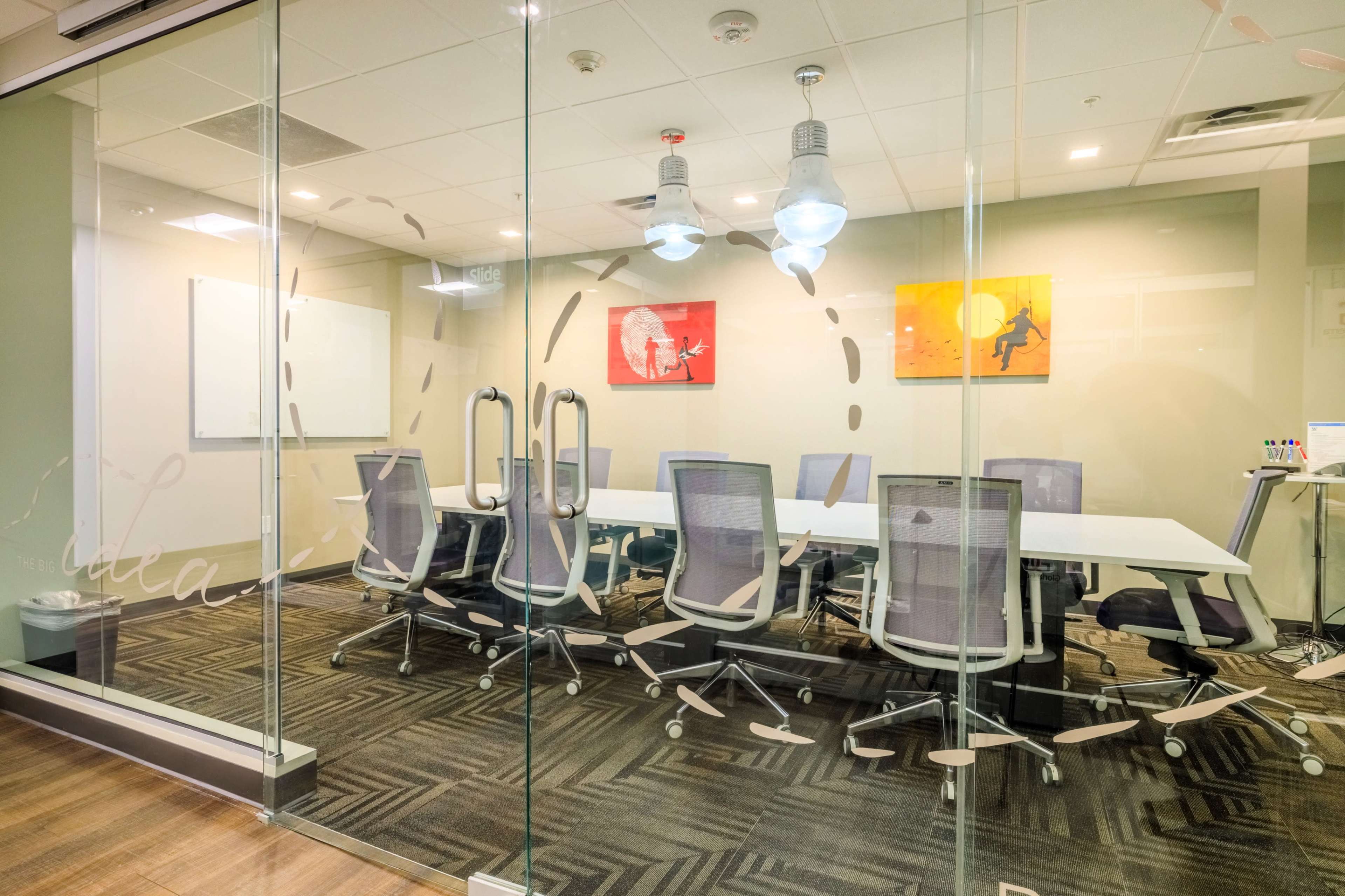 The image shows a modern conference room with a long white table surrounded by office chairs, enclosed by glass walls, and featuring artwork on the walls.