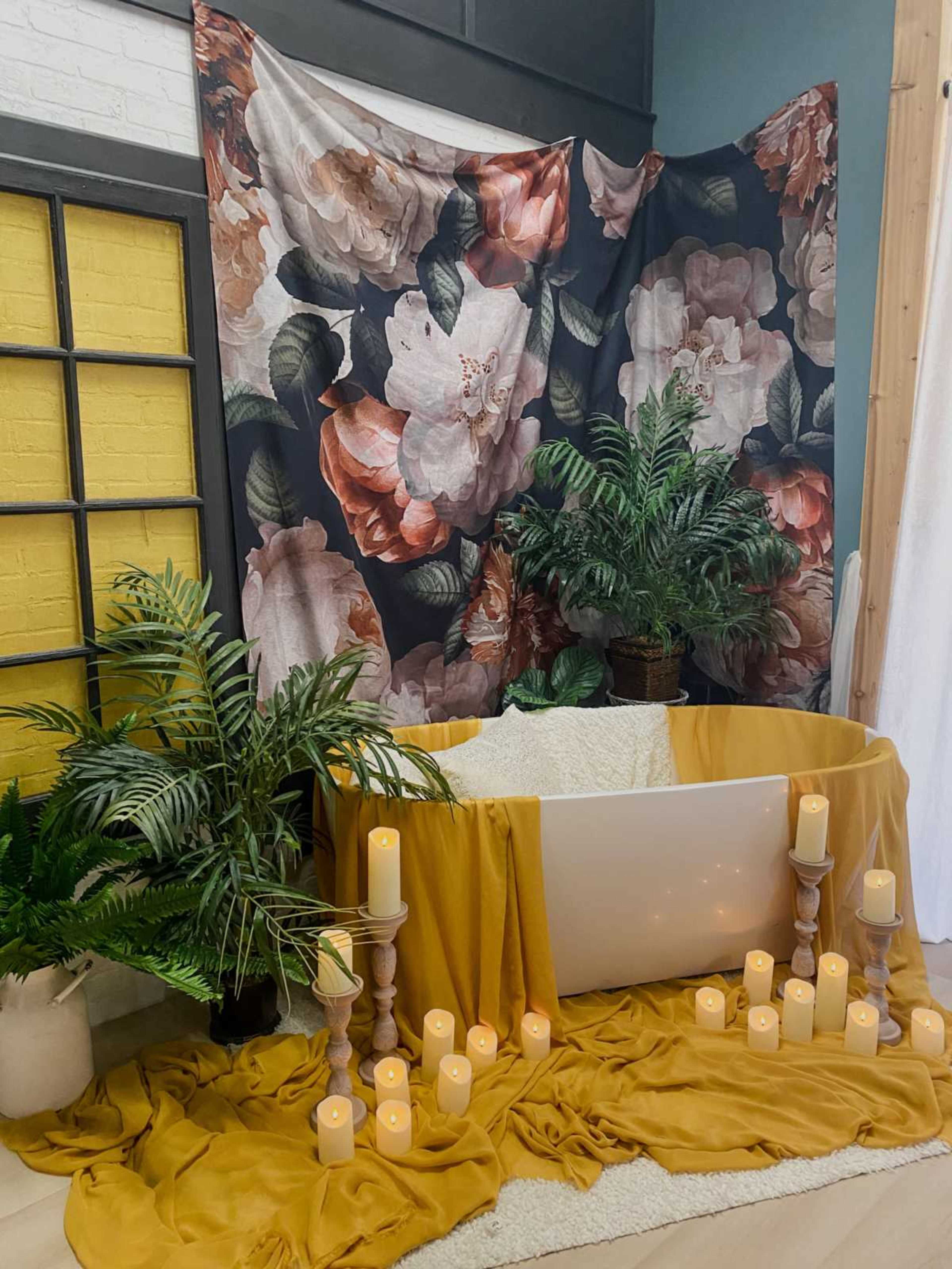 The image features a decorative space with a freestanding bathtub surrounded by yellow and white draping, lit candles, and lush greenery in the backdrop.