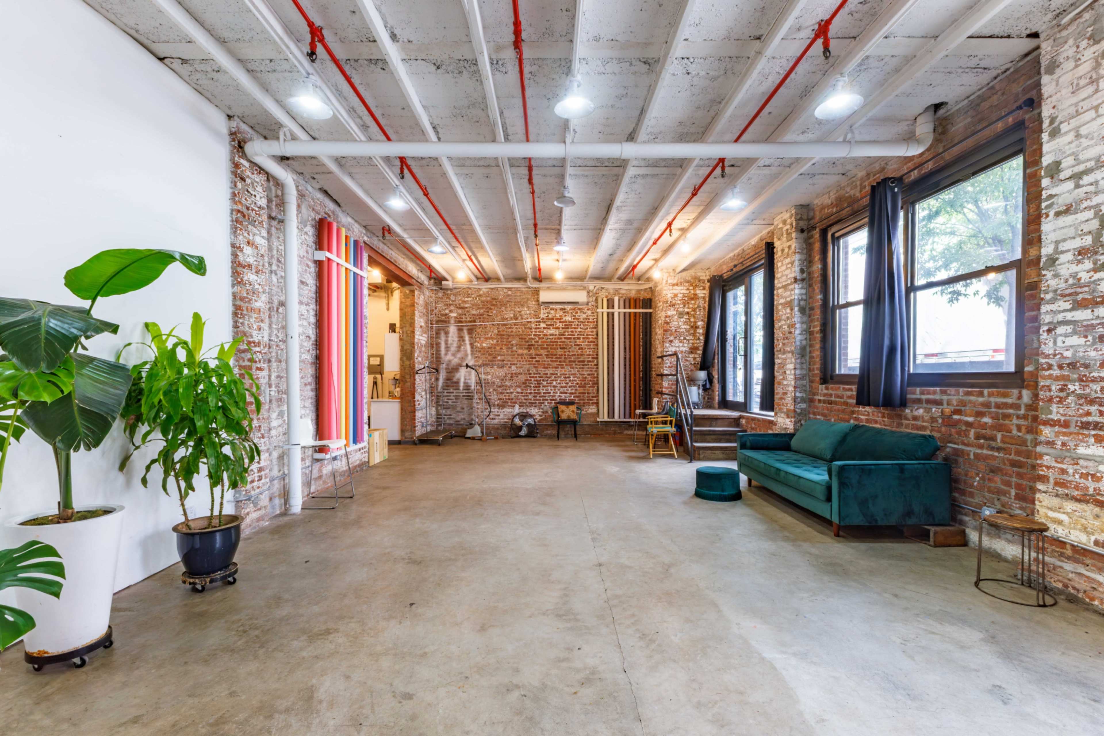 The image shows an industrial-style open space with exposed brick walls, concrete flooring, large windows, and a green sofa, along with plants and minimal furniture.