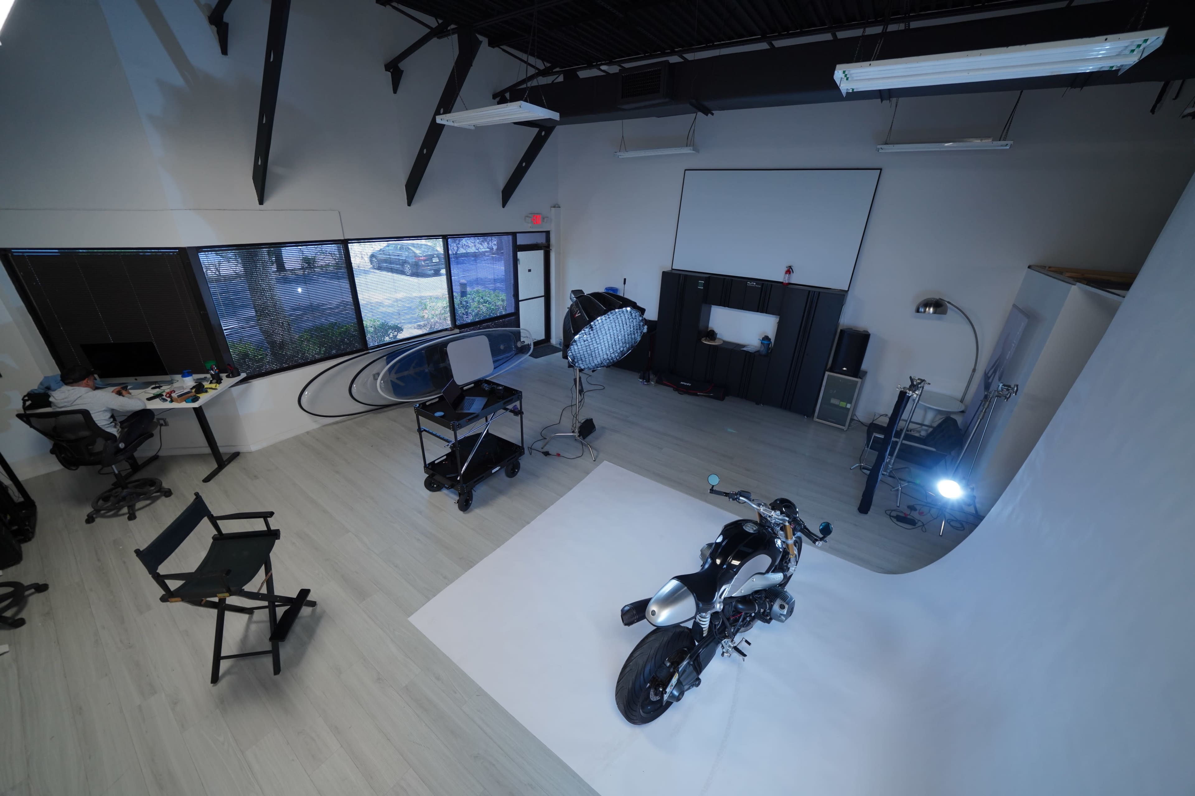 The image shows a spacious studio with a motorcycle placed on a white backdrop, surrounded by various equipment and large windows.