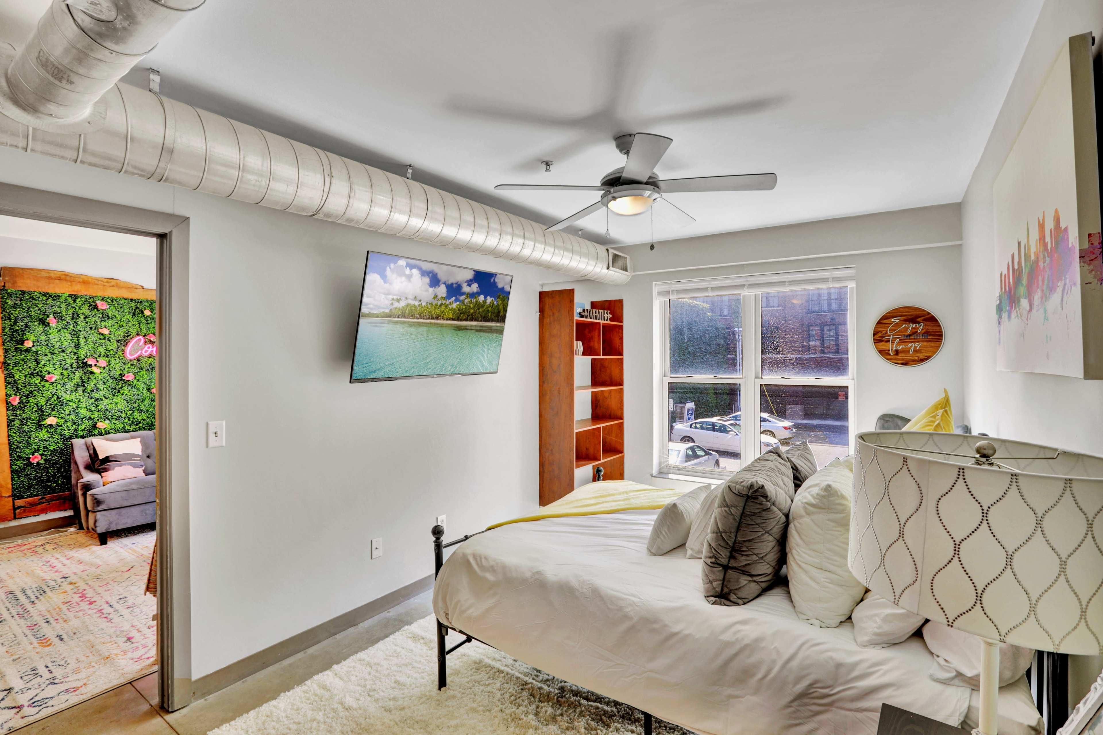 A compact bedroom featuring a bed with decorative pillows, a ceiling fan, a television mounted on the wall, and a window revealing an outdoor view.