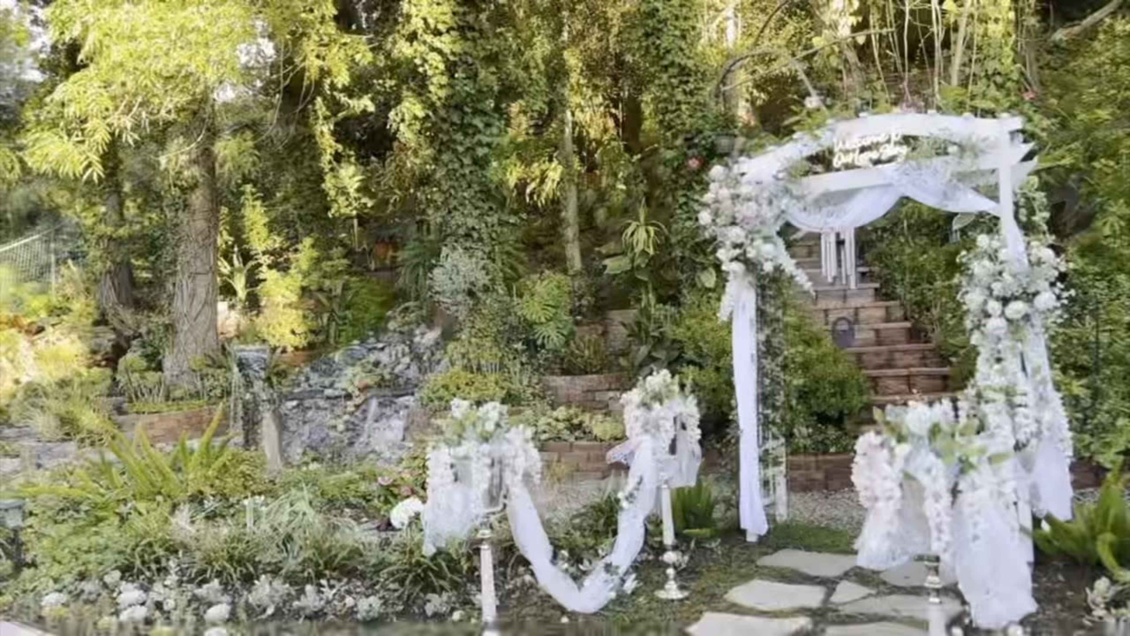 The image features a beautifully decorated outdoor wedding setting with an arch adorned with flowers and white fabric, surrounded by lush greenery and a path leading to stone steps.