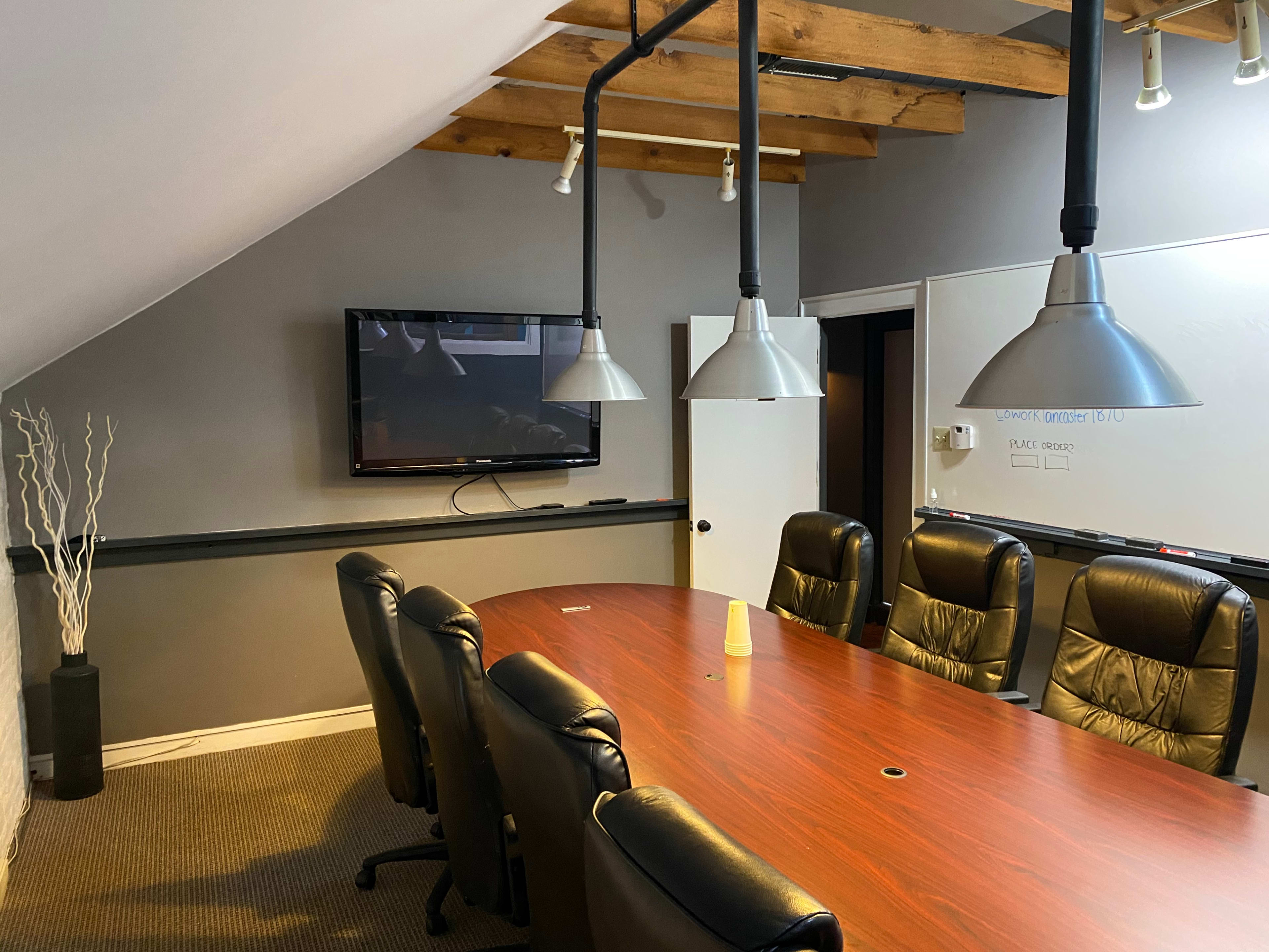 A conference room features a long wooden table surrounded by black leather chairs, with a television mounted on the wall and two large pendant lights above.