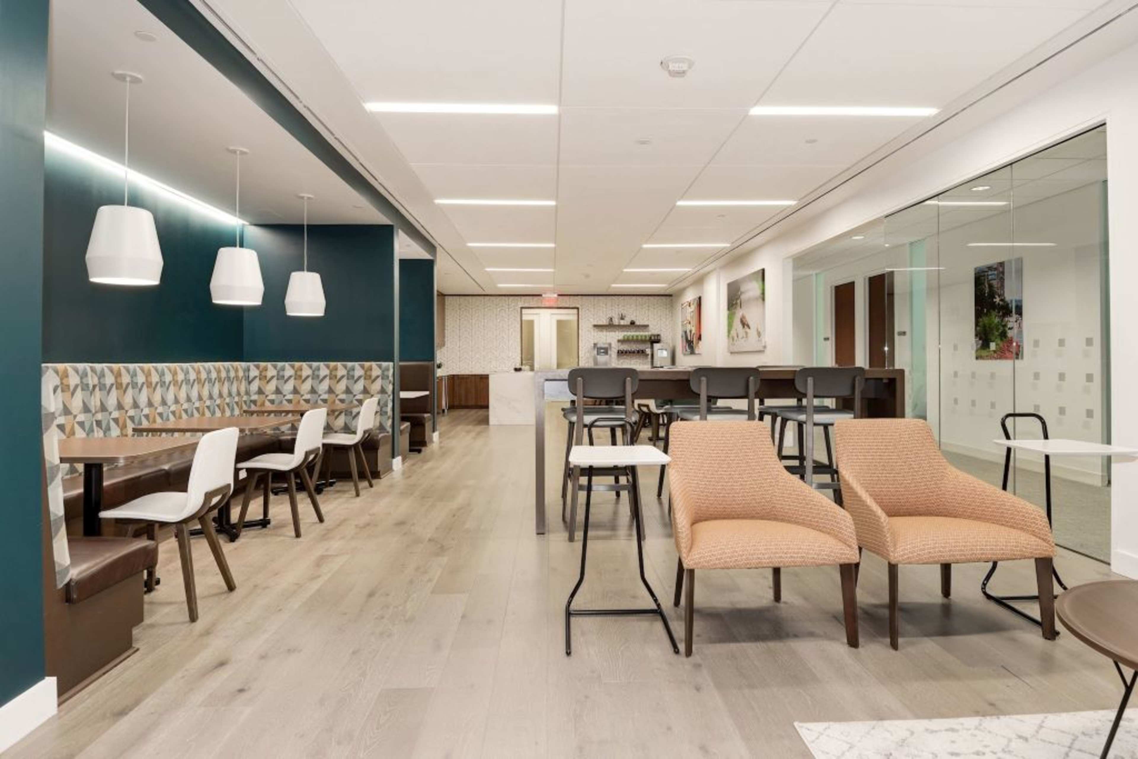 A modern office lounge area with tables, chairs, and a kitchenette in a well-lit space.