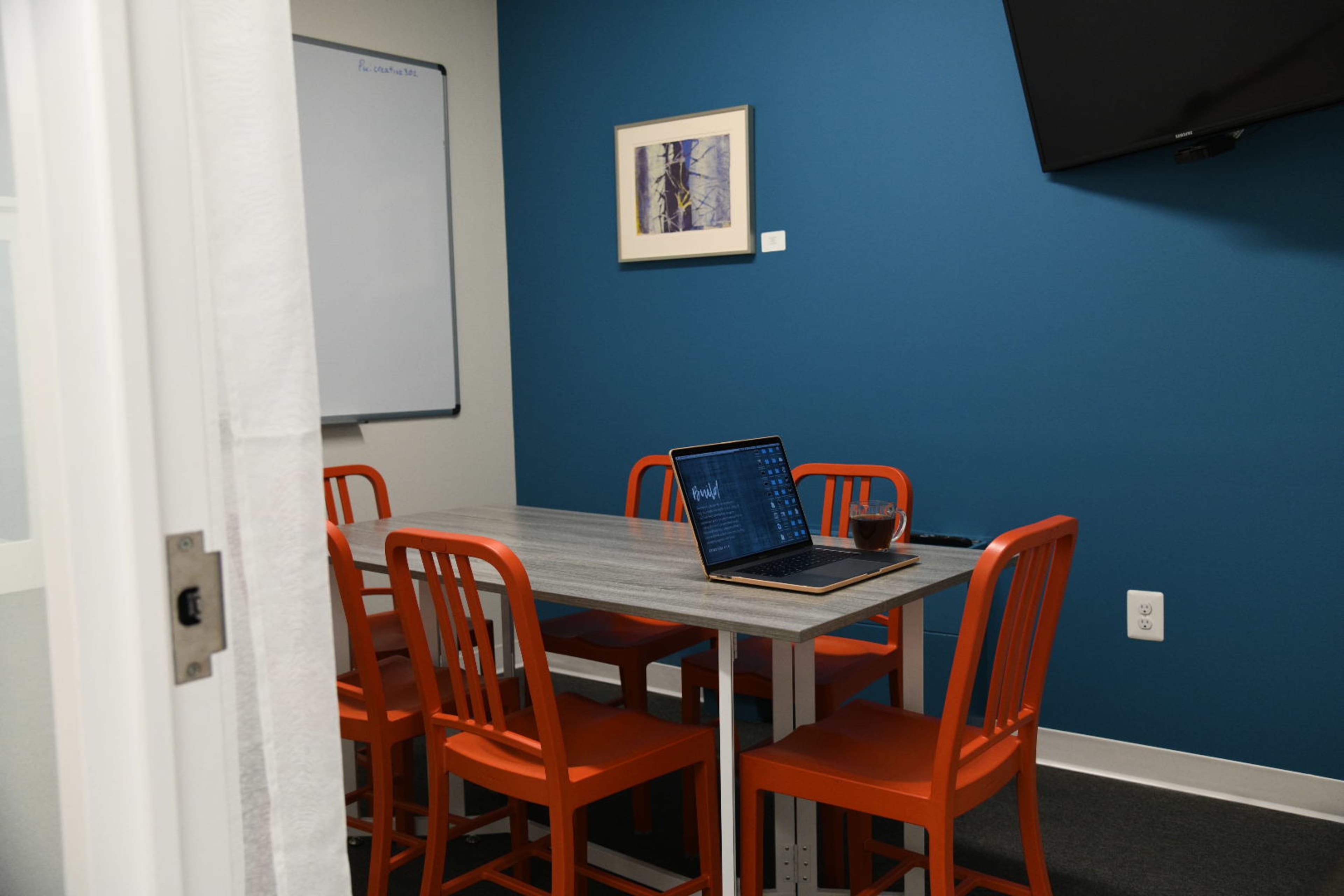 A conference room features a wooden table with a laptop and a glass sitting on it, surrounded by orange chairs against a blue wall.