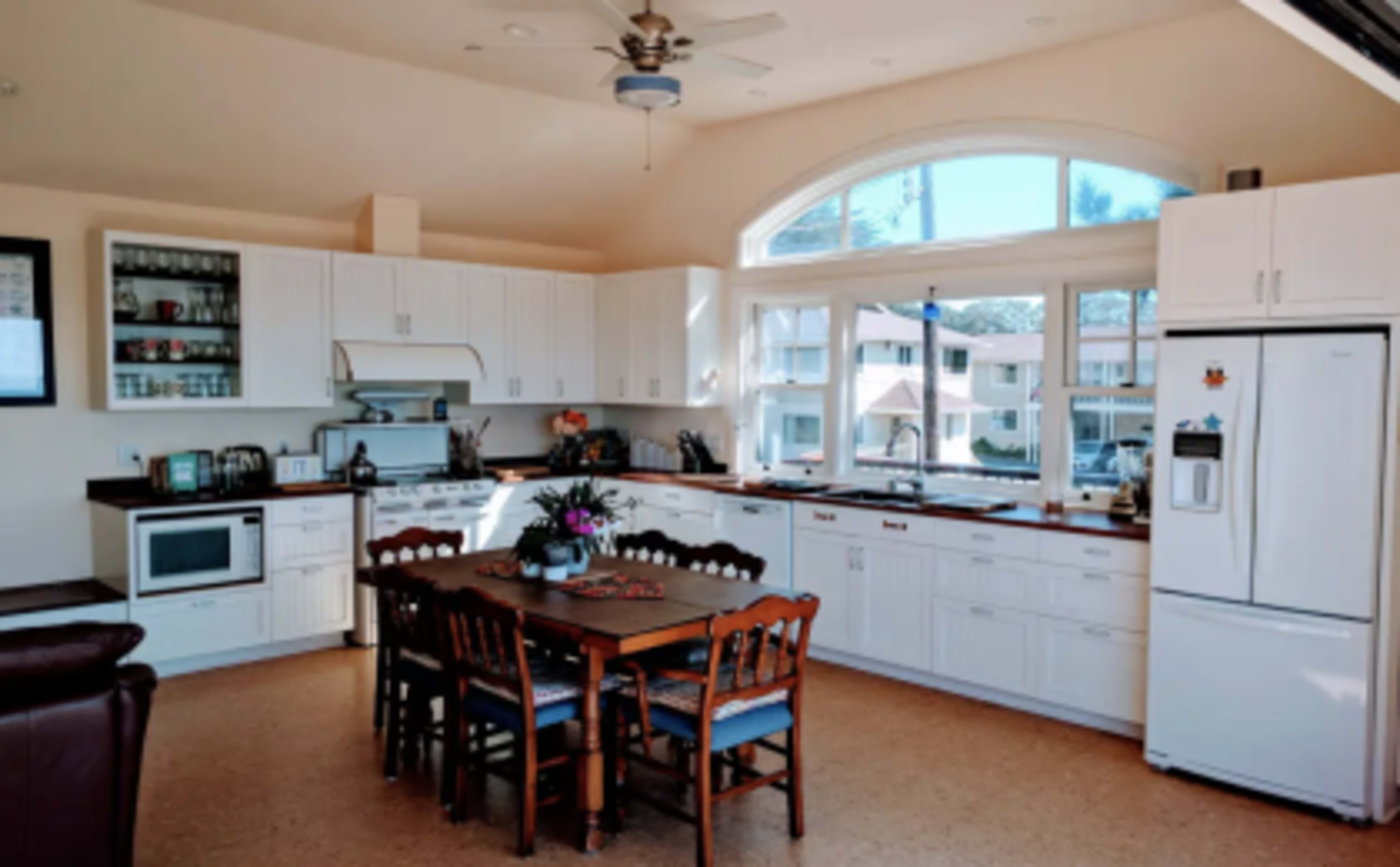 The image shows a spacious kitchen with white cabinetry, a large table surrounded by chairs, and a window that lets in natural light.