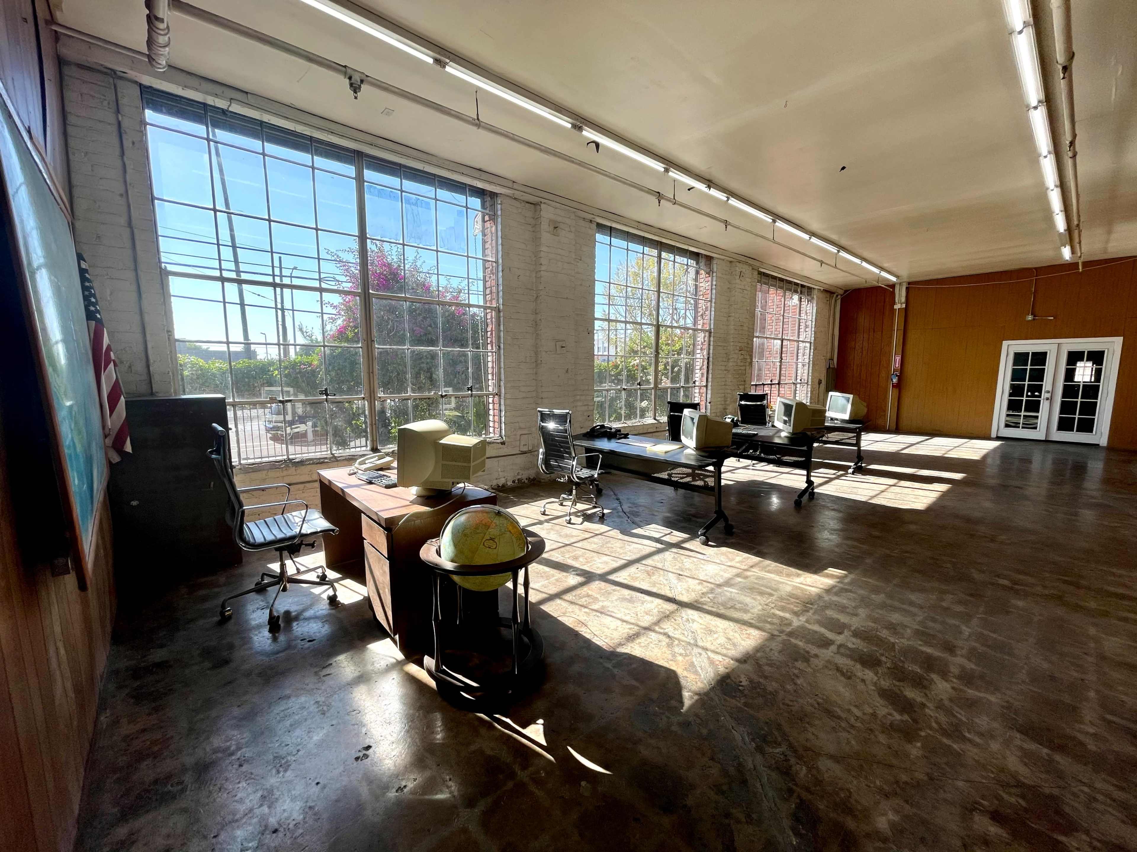 The image shows a spacious, well-lit room with large windows, featuring several desks and chairs, and a globe on a table near the foreground.