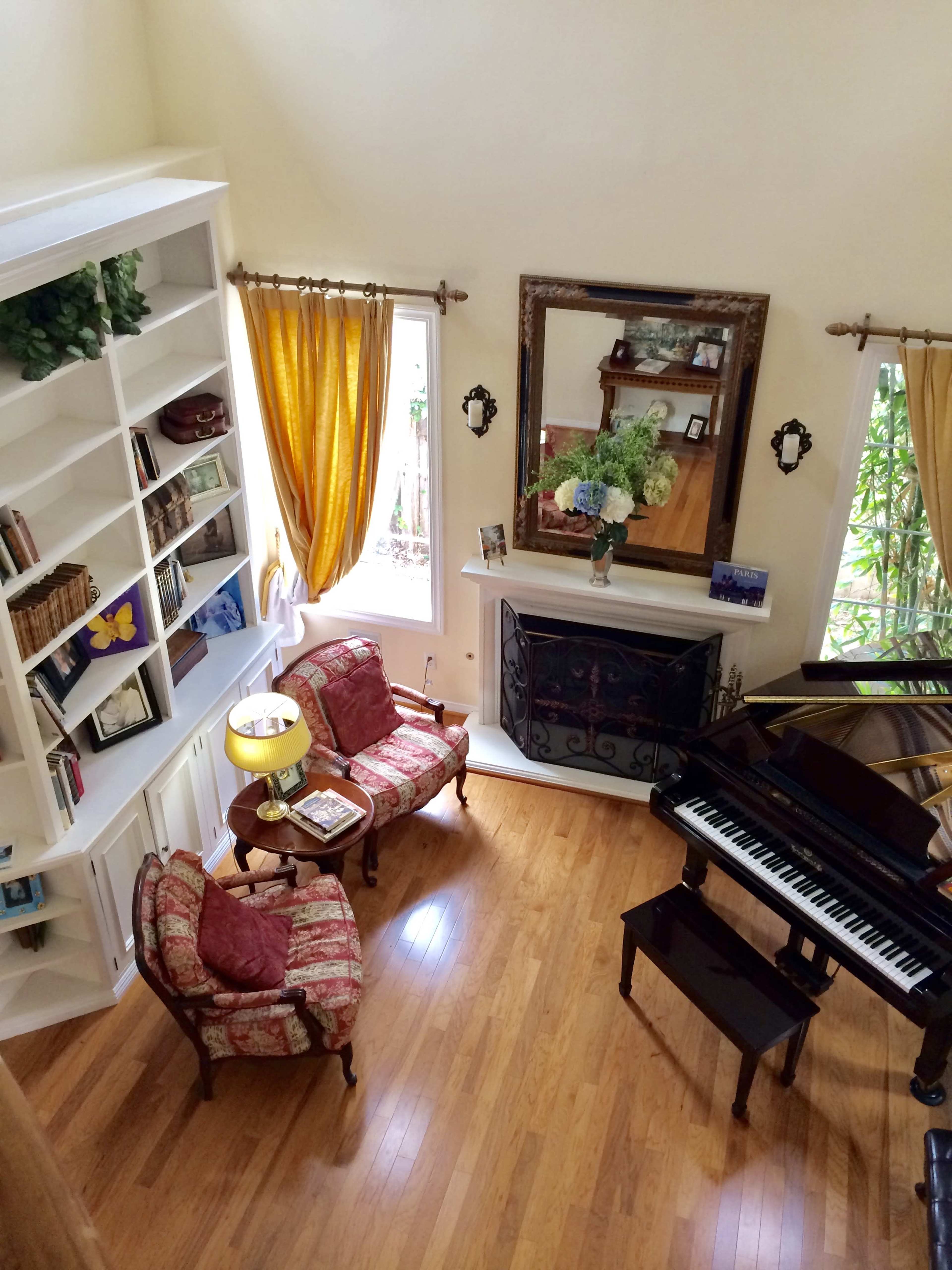 A living room features two chairs, a lamp, a piano, a fireplace, and a bookshelf, all arranged around large windows with yellow curtains.