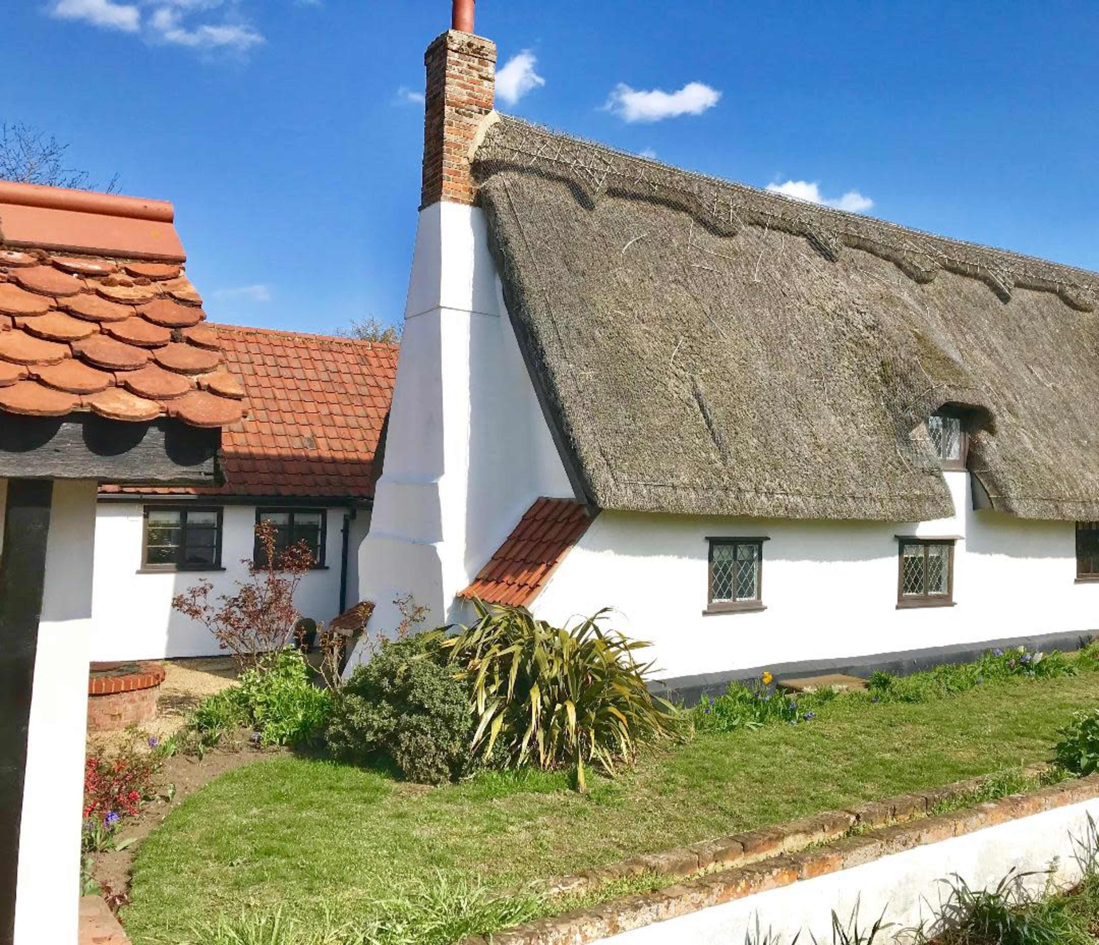 A thatched cottage with a brick chimney is surrounded by a grassy garden and flowering plants.