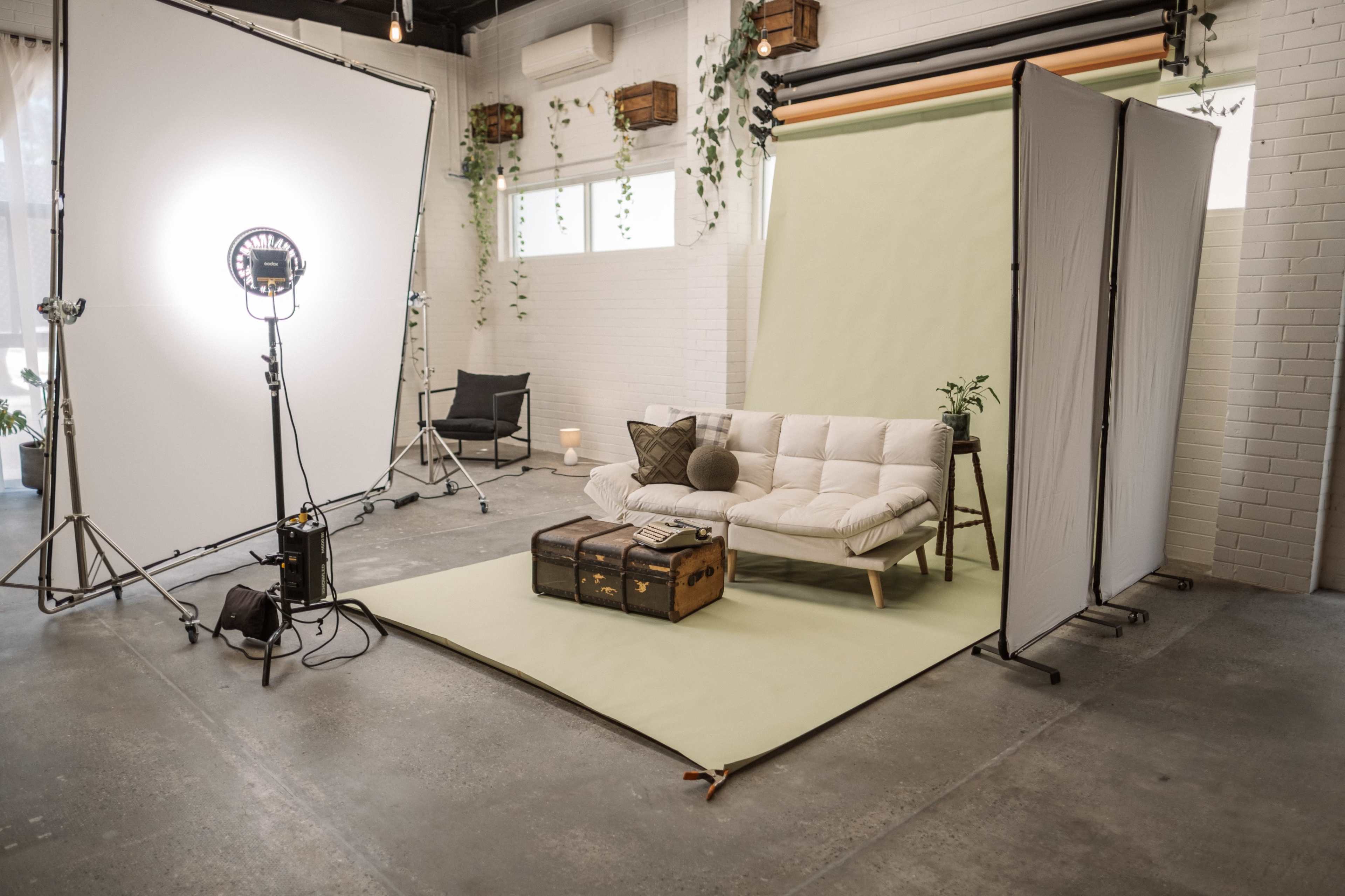 A modern studio setup featuring a green backdrop, a light source, a white sofa with decorative pillows, a vintage trunk, and various plants.