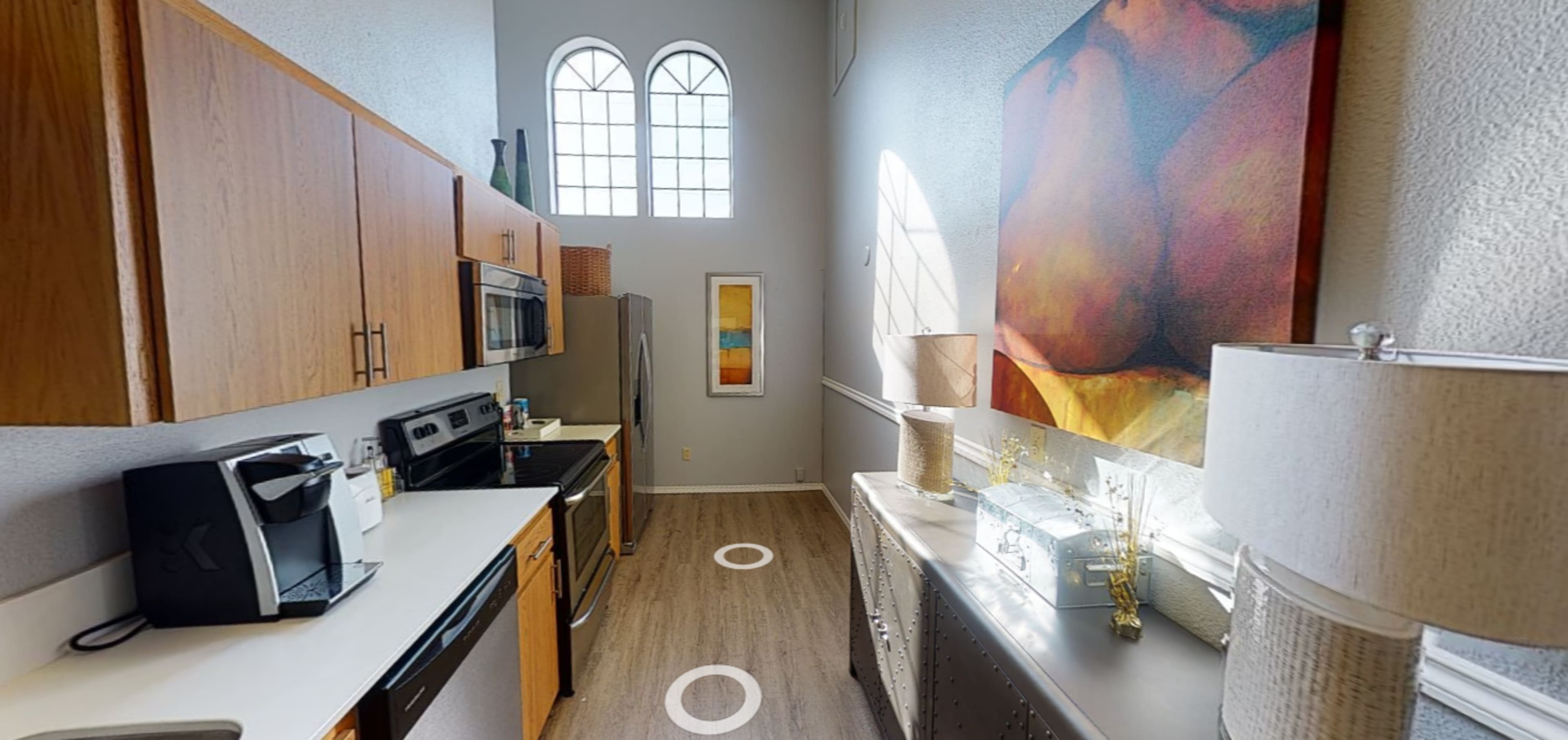 A kitchen with wooden cabinets, stainless steel appliances, and a large artwork on the wall, illuminated by natural light from arched windows.