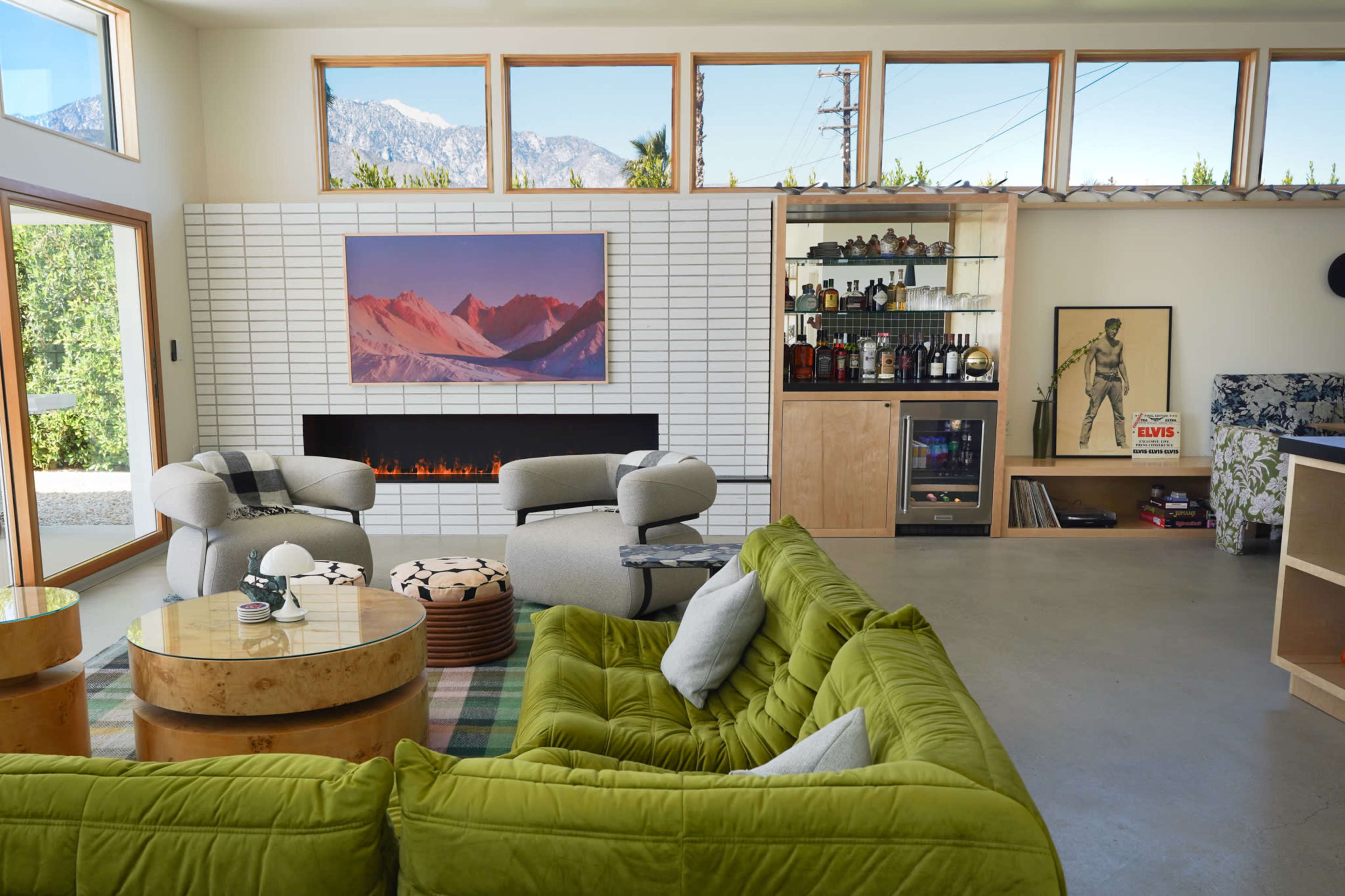 A modern living room with a green sofa, a coffee table, a wall-mounted fireplace, and a bar area, all framed by large windows that reveal a view of mountains outside.