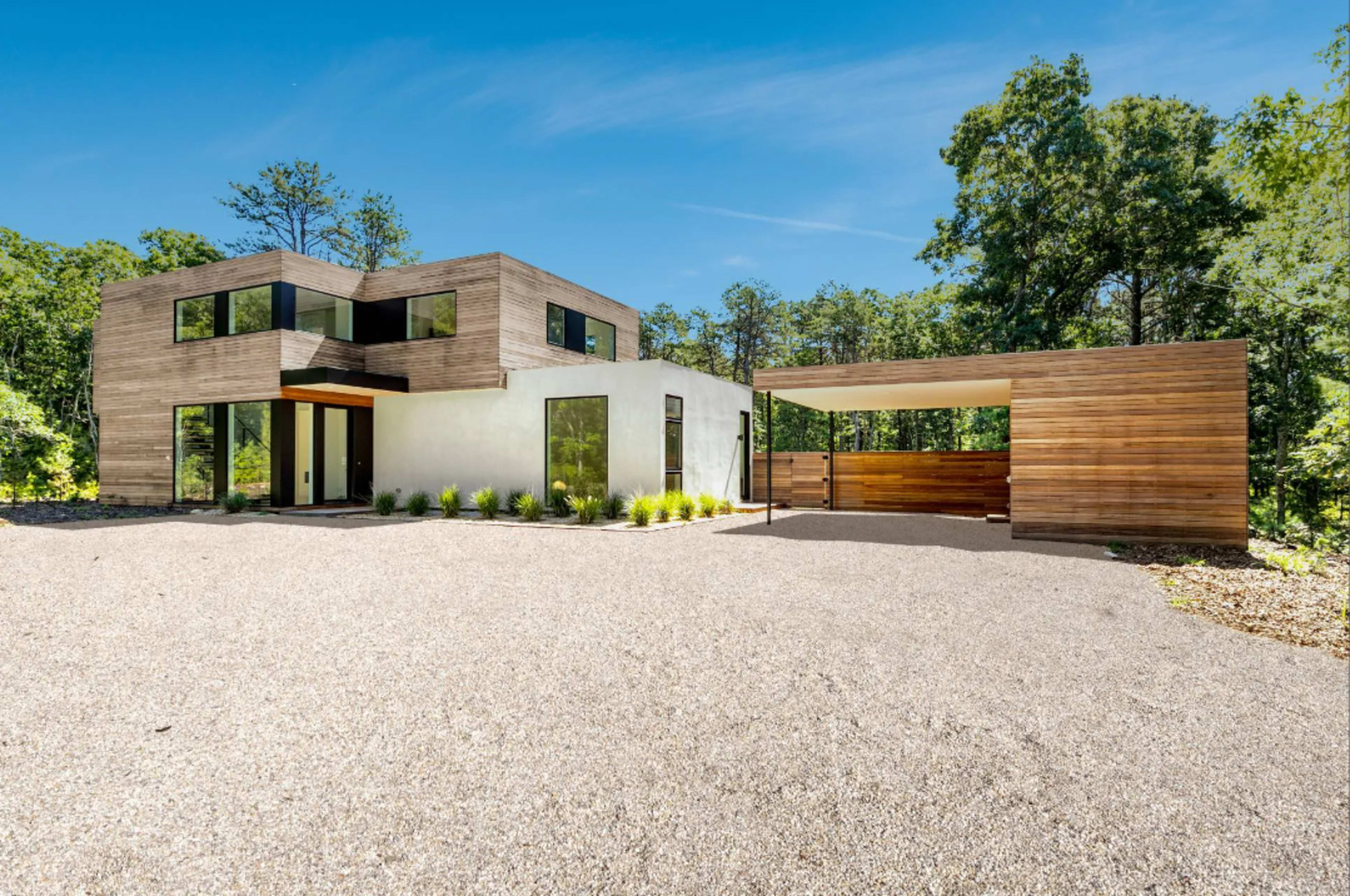 A modern two-story house with a flat roof and a wooden exterior is situated in a wooded area, accompanied by a carport.