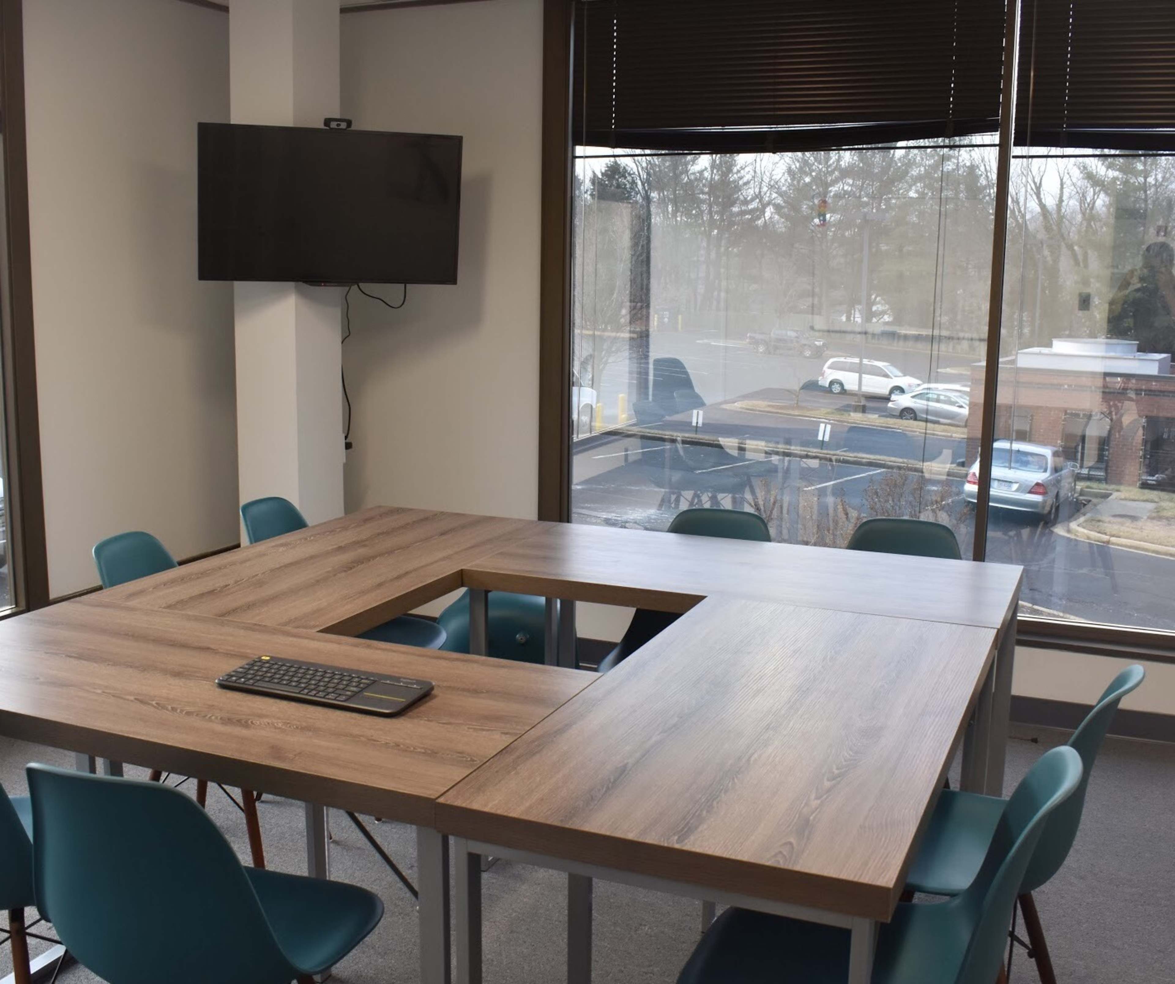 A modern conference room features a large table with a cutout in the center, surrounded by teal chairs, and a wall-mounted television.