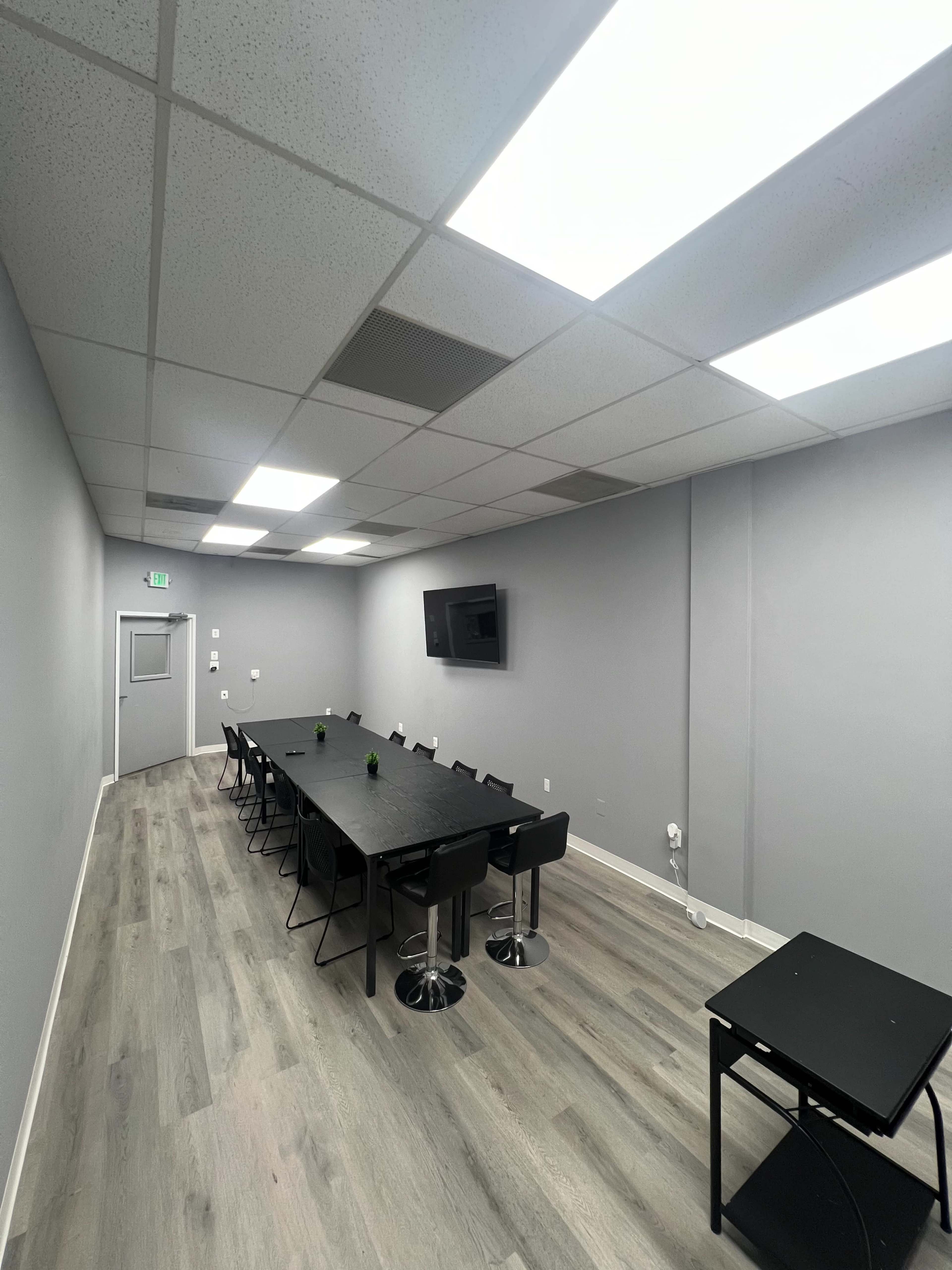 A conference room with a long black table surrounded by black chairs, positioned under fluorescent ceiling lights, with a television mounted on the wall.