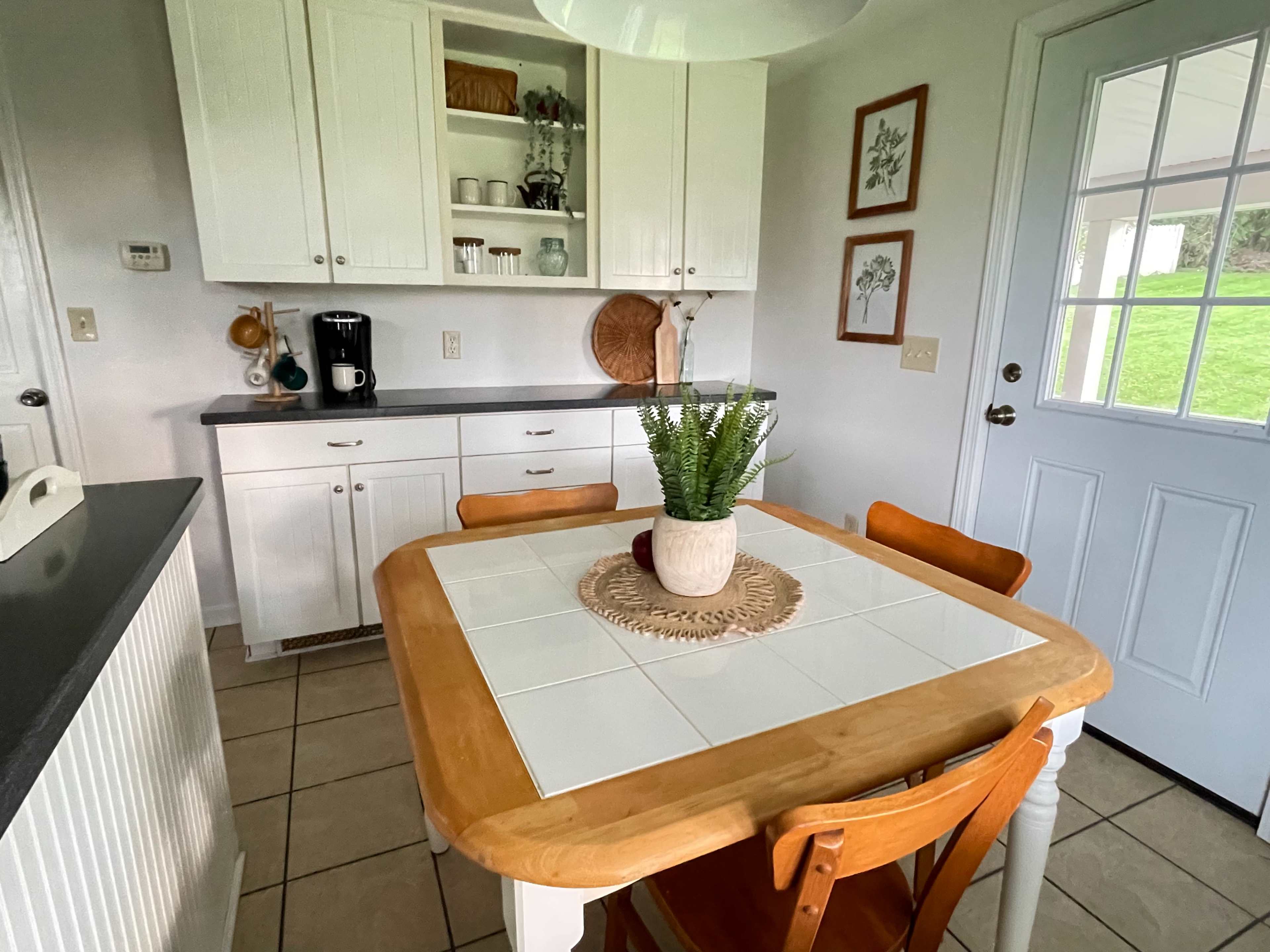 A bright kitchen features a wooden table with a tile centerpiece, surrounded by four wooden chairs, with white cabinets and a door leading outside.
