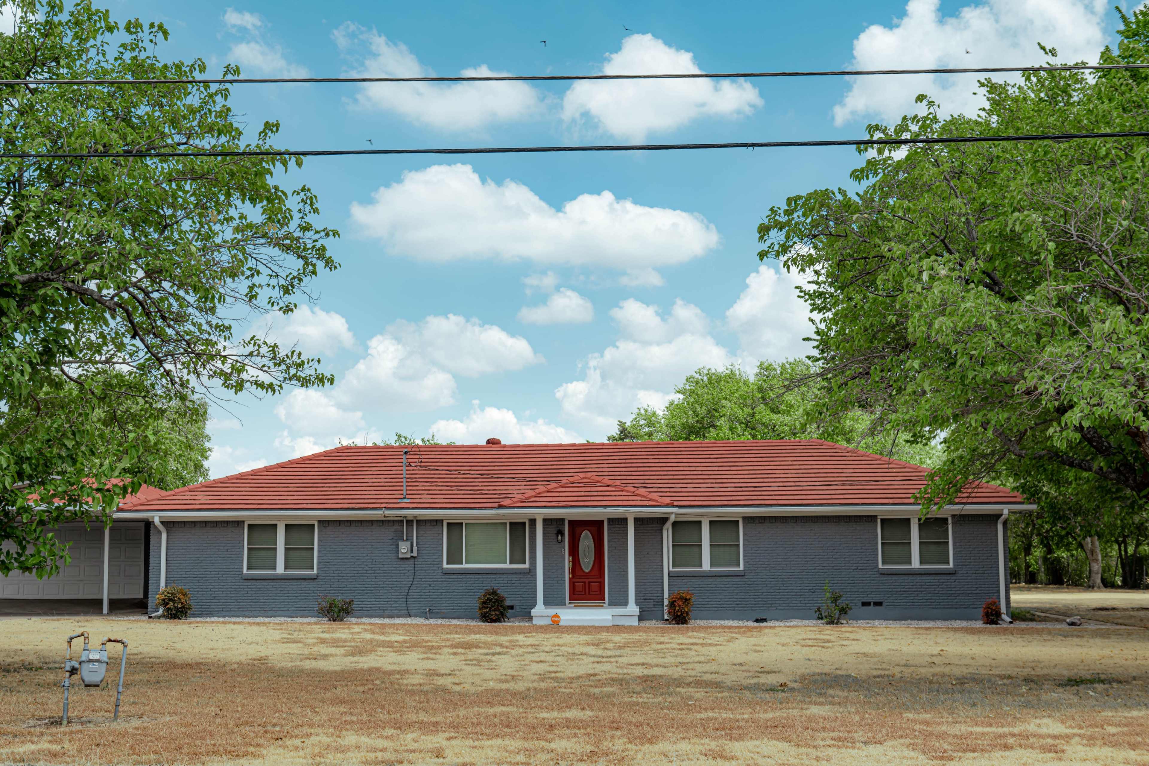 A single-story house with a red roof and gray exterior sits on a dry lawn under a blue sky with fluffy clouds.
