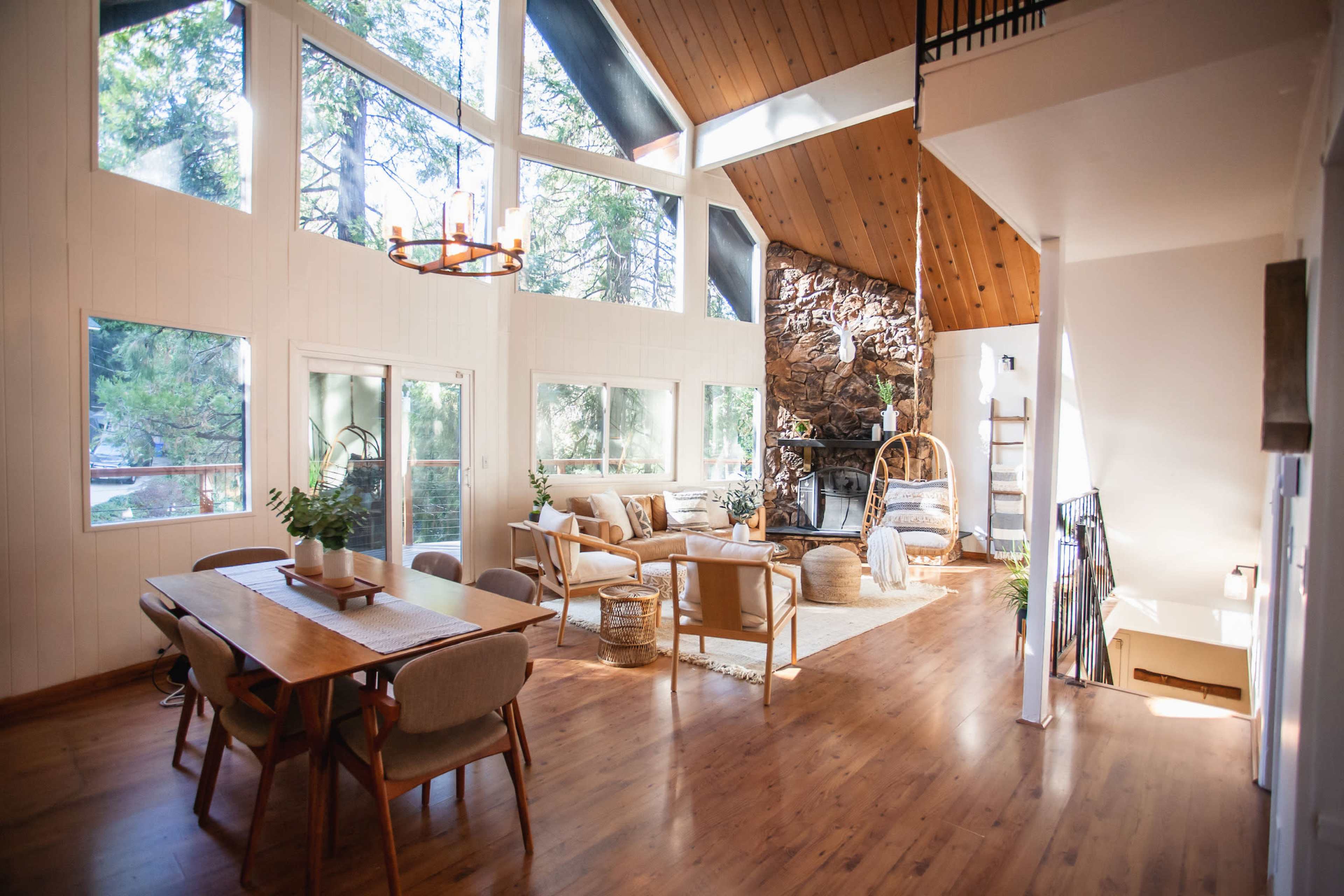 A bright, open living area with large windows, a stone fireplace, and a dining table, surrounded by natural wood finishes and furniture.