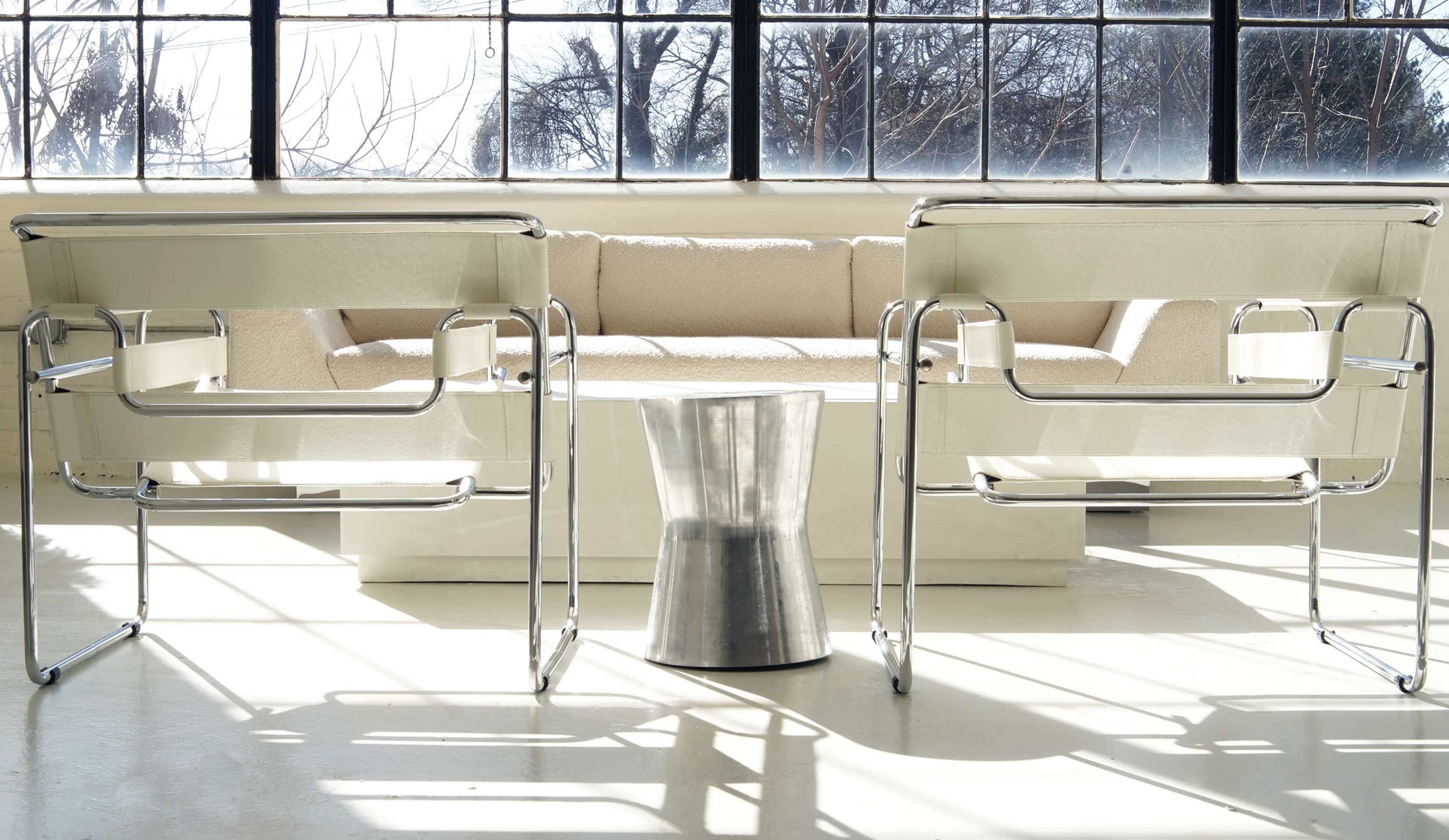 Two metal-framed chairs facing a light-colored sofa with a metallic side table in between, all set against large windows letting in natural light.