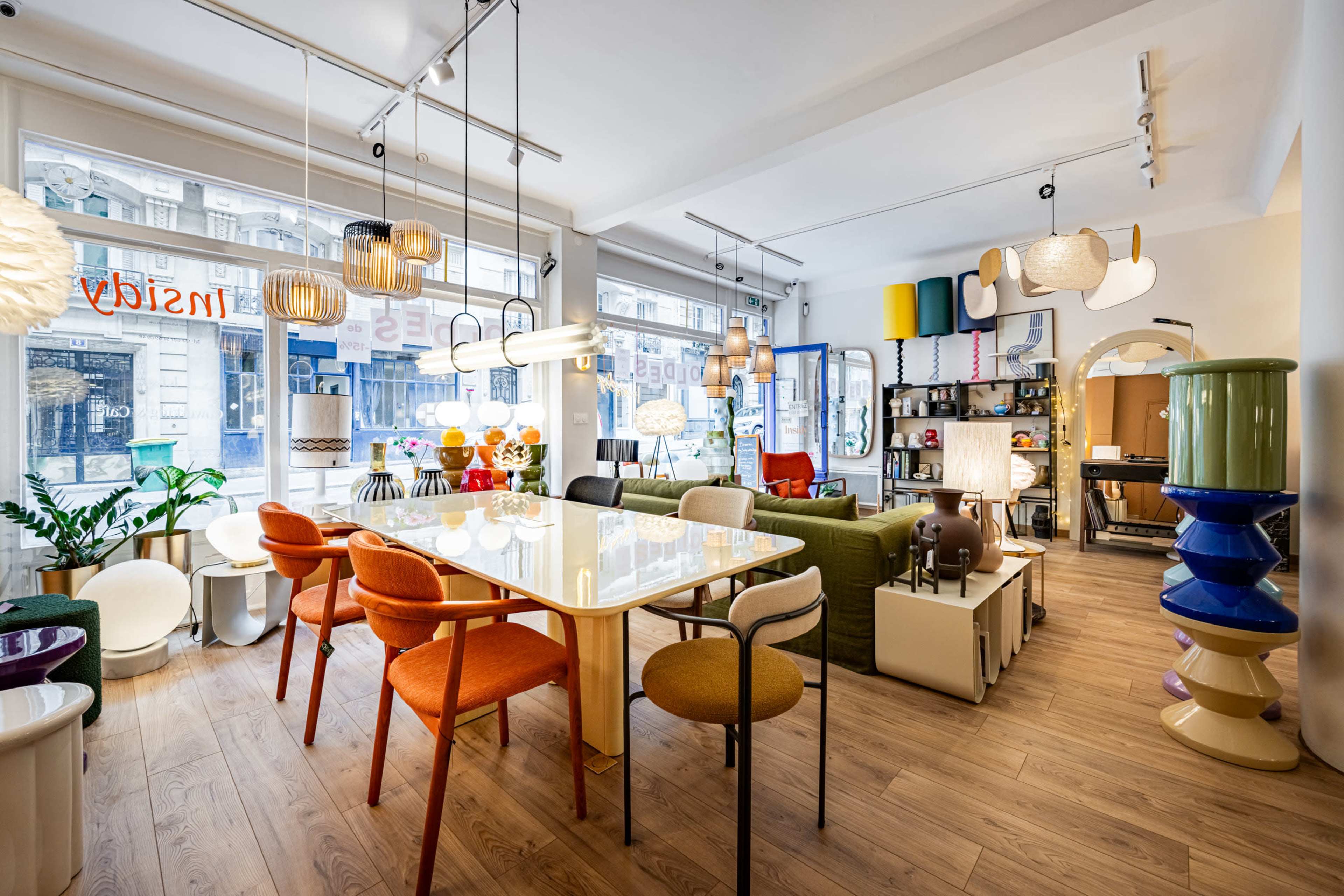 The image shows a modern furniture showroom featuring a variety of colorful chairs, a large dining table, and decorative light fixtures, with large windows allowing natural light to enter.