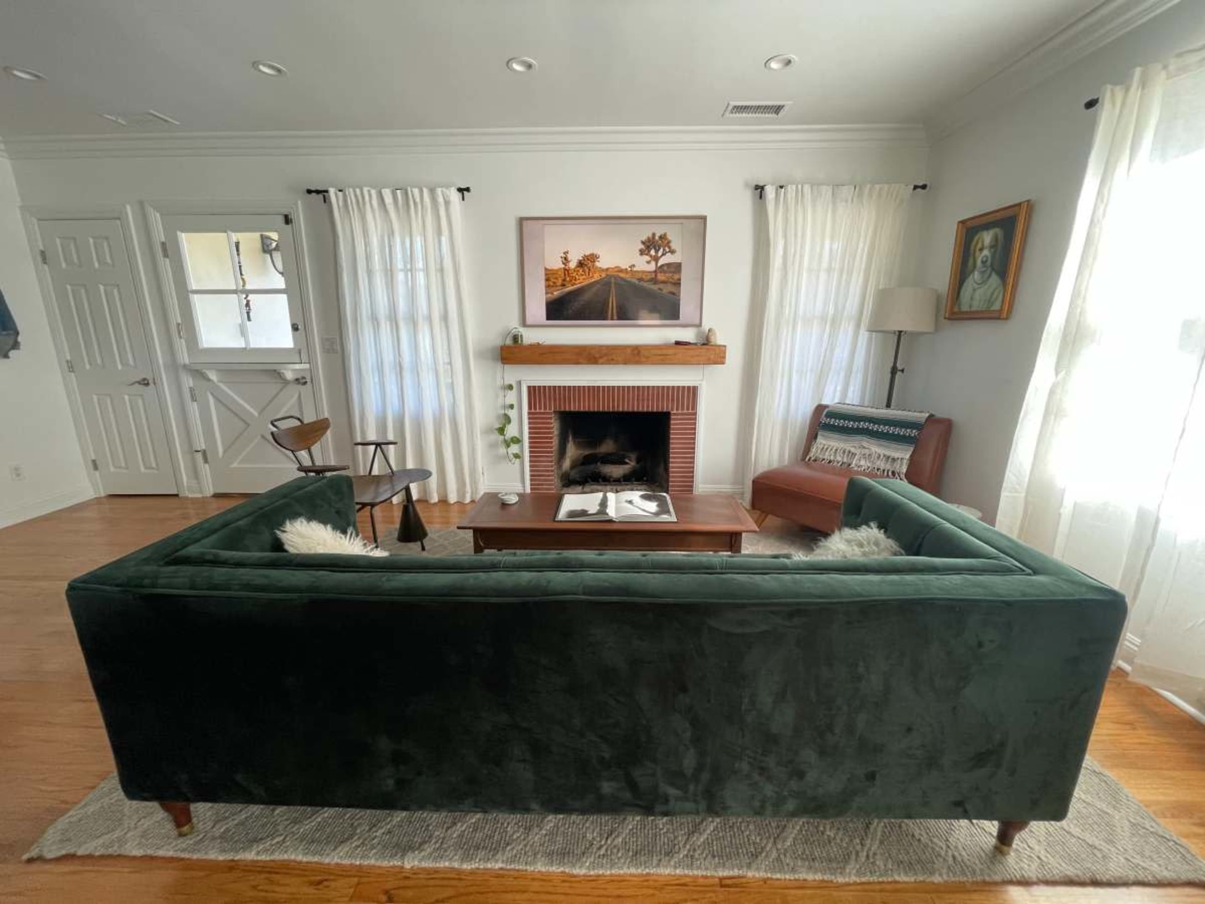 The image shows a cozy living room featuring a green sectional sofa, a wooden coffee table, a fireplace, and natural light streaming through sheer curtains.