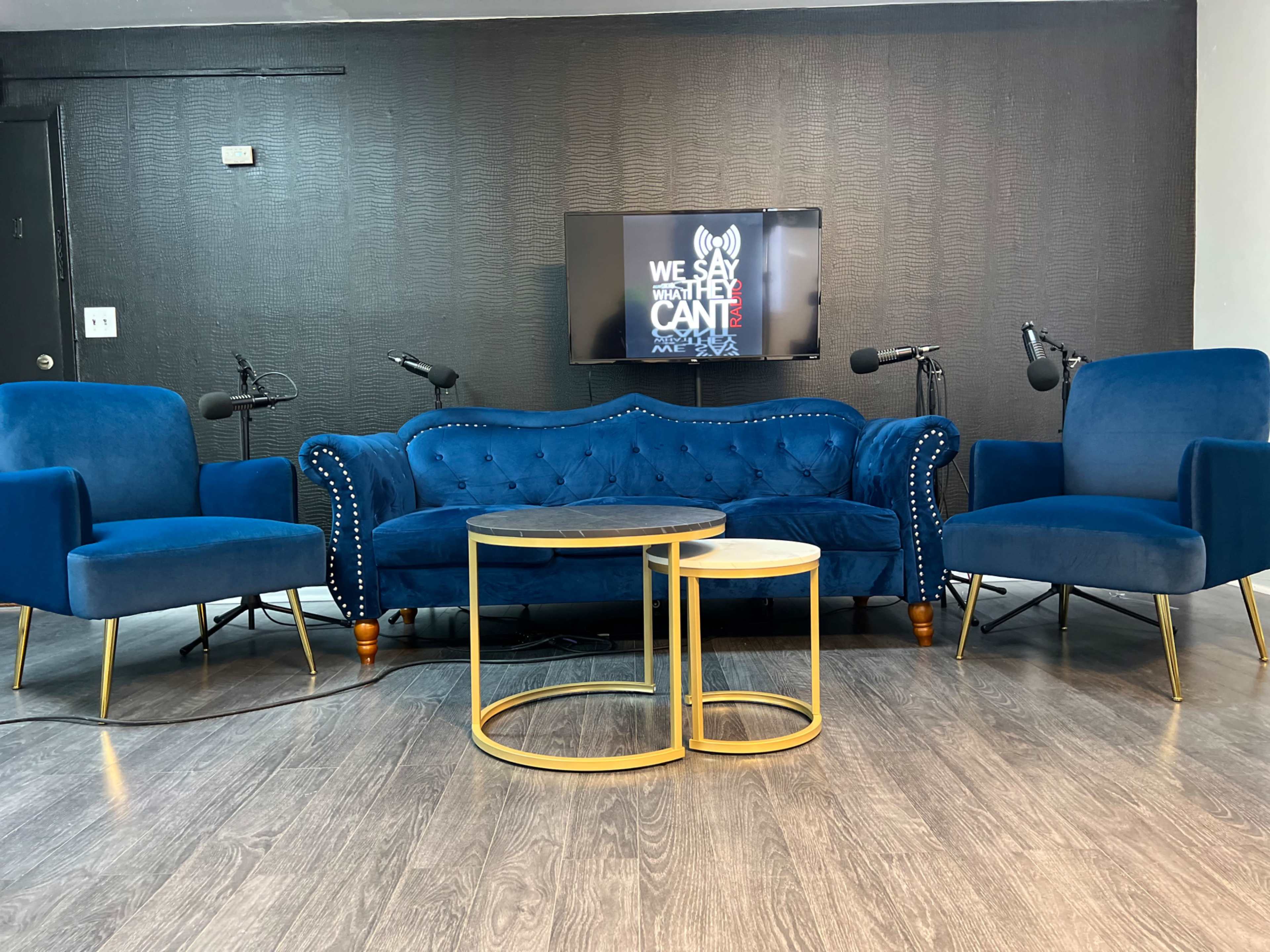The image shows a modern seating area with a blue velvet couch and two matching armchairs, accompanied by a round coffee table, with microphones set up for a podcast recording in front of a wall-mounted television.