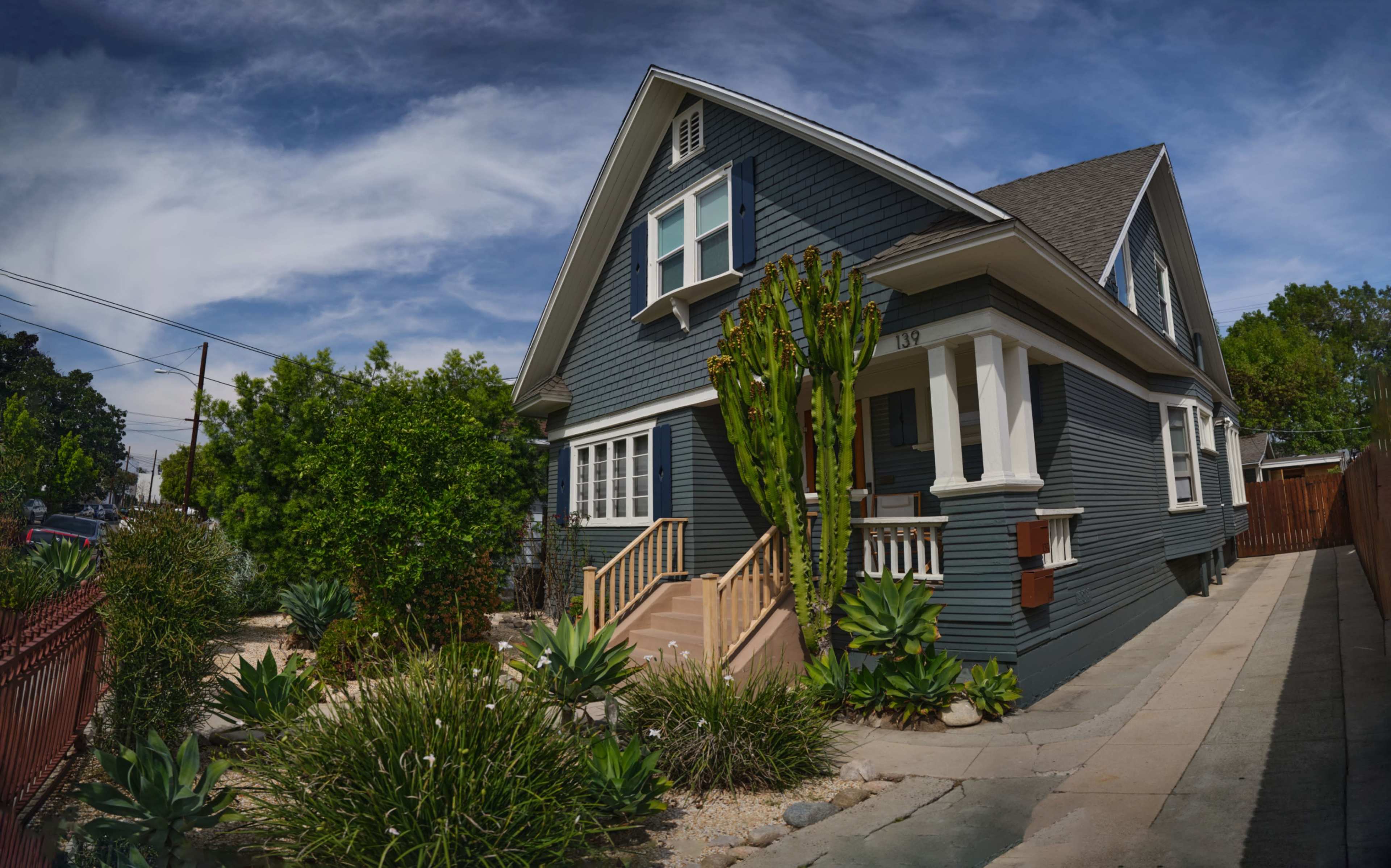 The image shows a two-story blue-gray house with a prominent front porch, surrounded by desert plants and a landscaped yard, under a partly cloudy sky.
