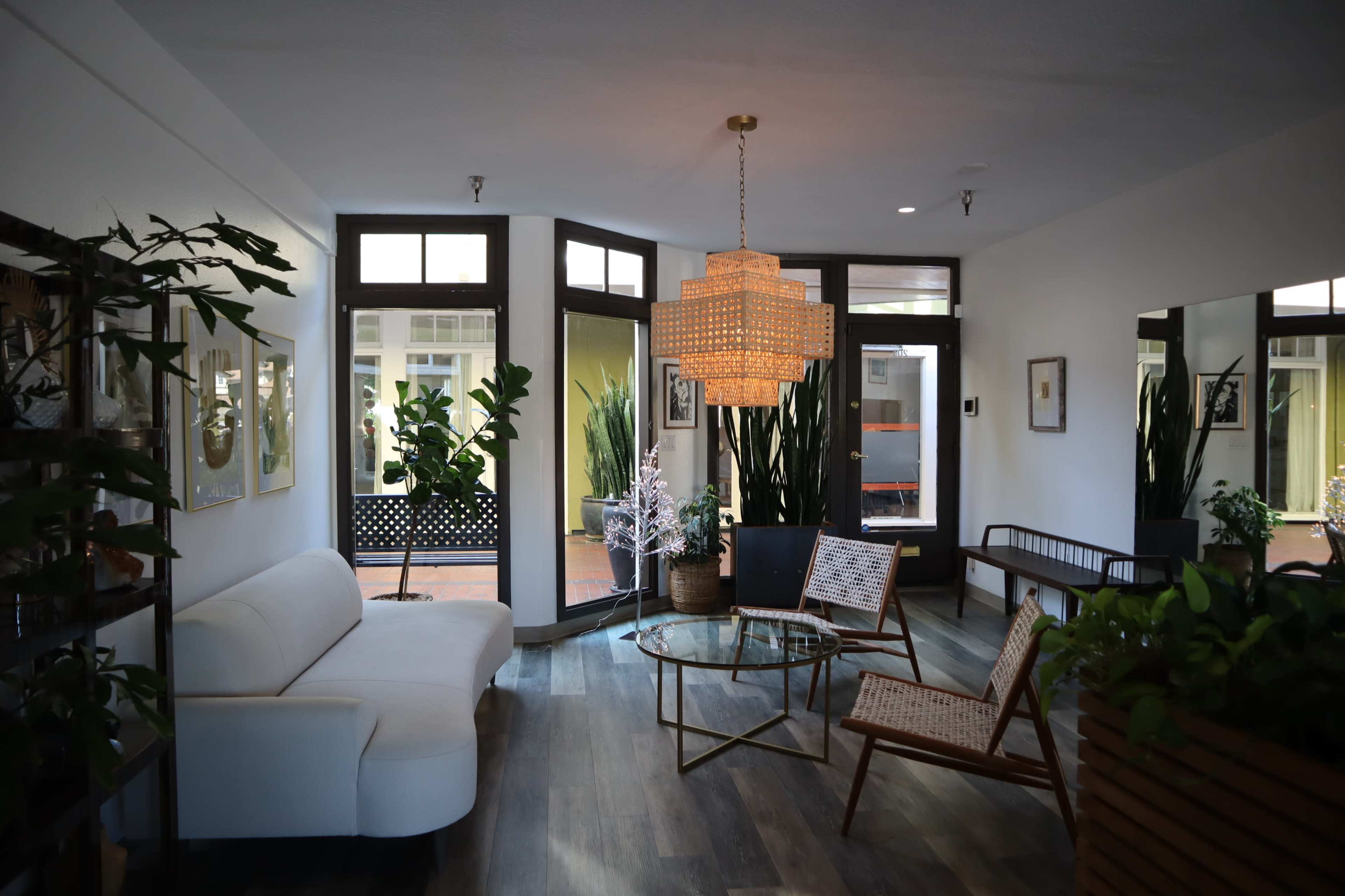 A modern lounge area featuring a white sofa, a round glass coffee table, wicker chairs, and large indoor plants, with ample natural light coming through the windows.