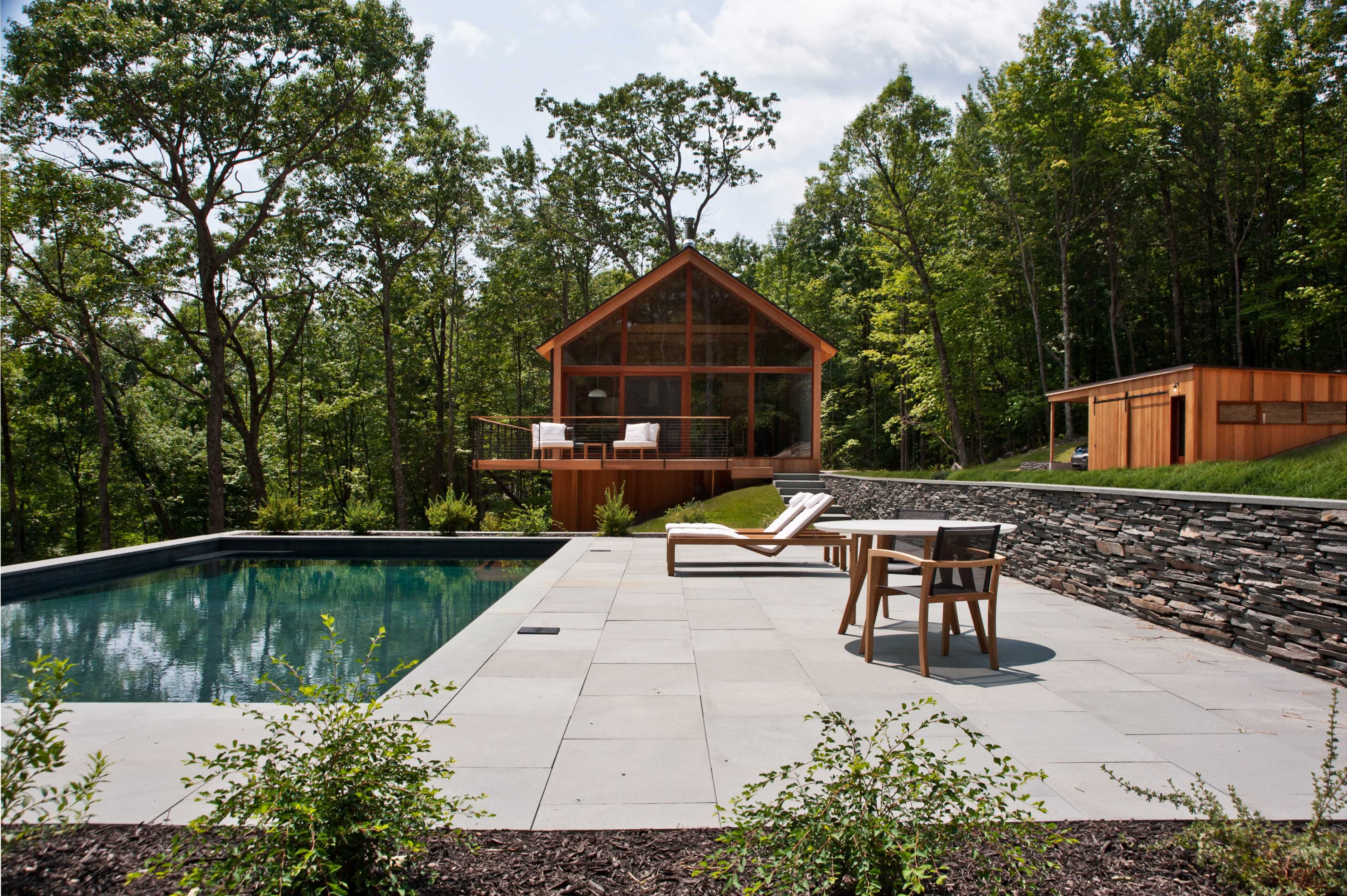 A modern house with large windows overlooking a rectangular swimming pool, surrounded by a stone patio and greenery.