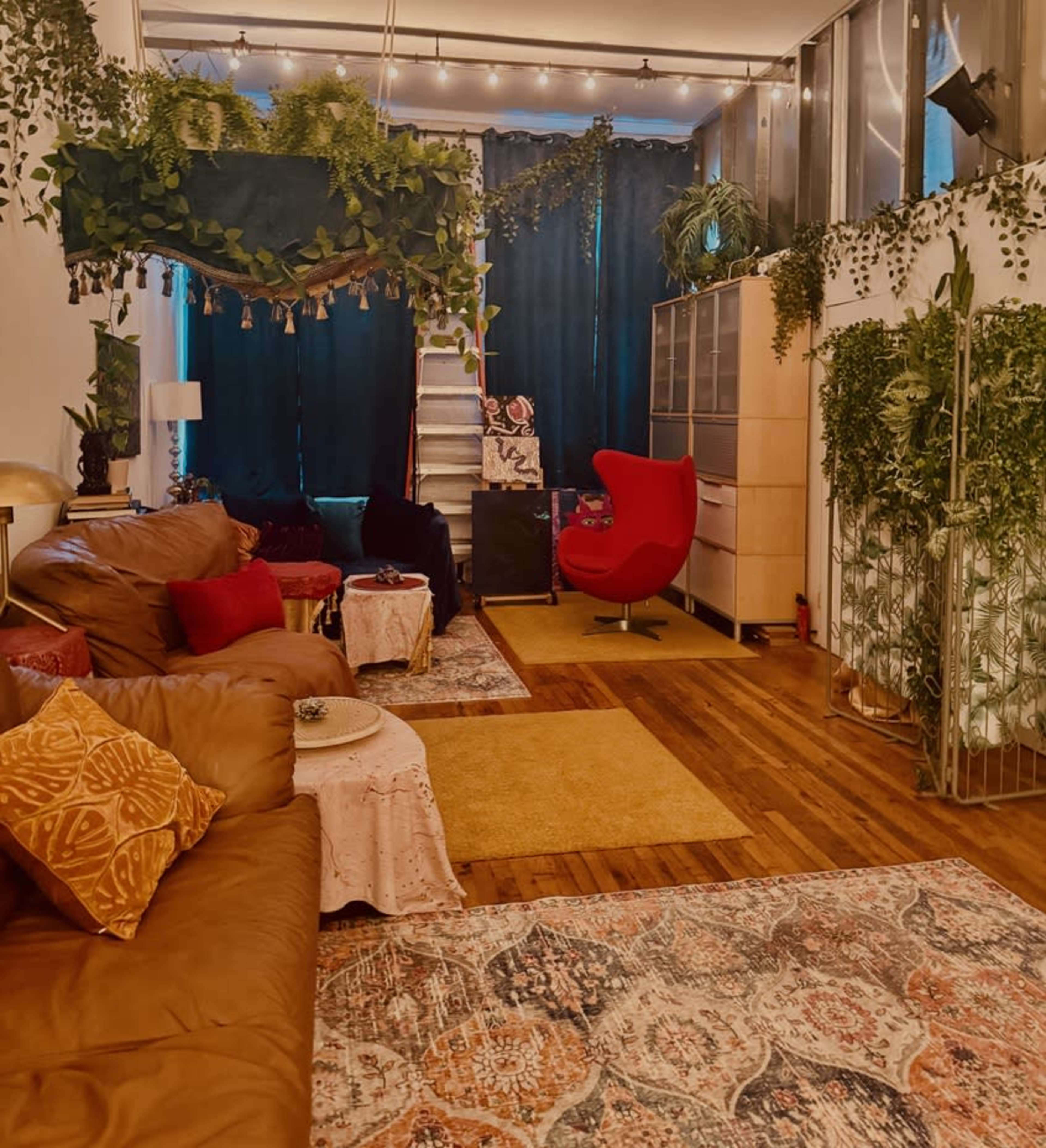 The image shows a cozy living space with a brown leather couch, colorful rugs, potted plants hanging from the ceiling, and a red accent chair.