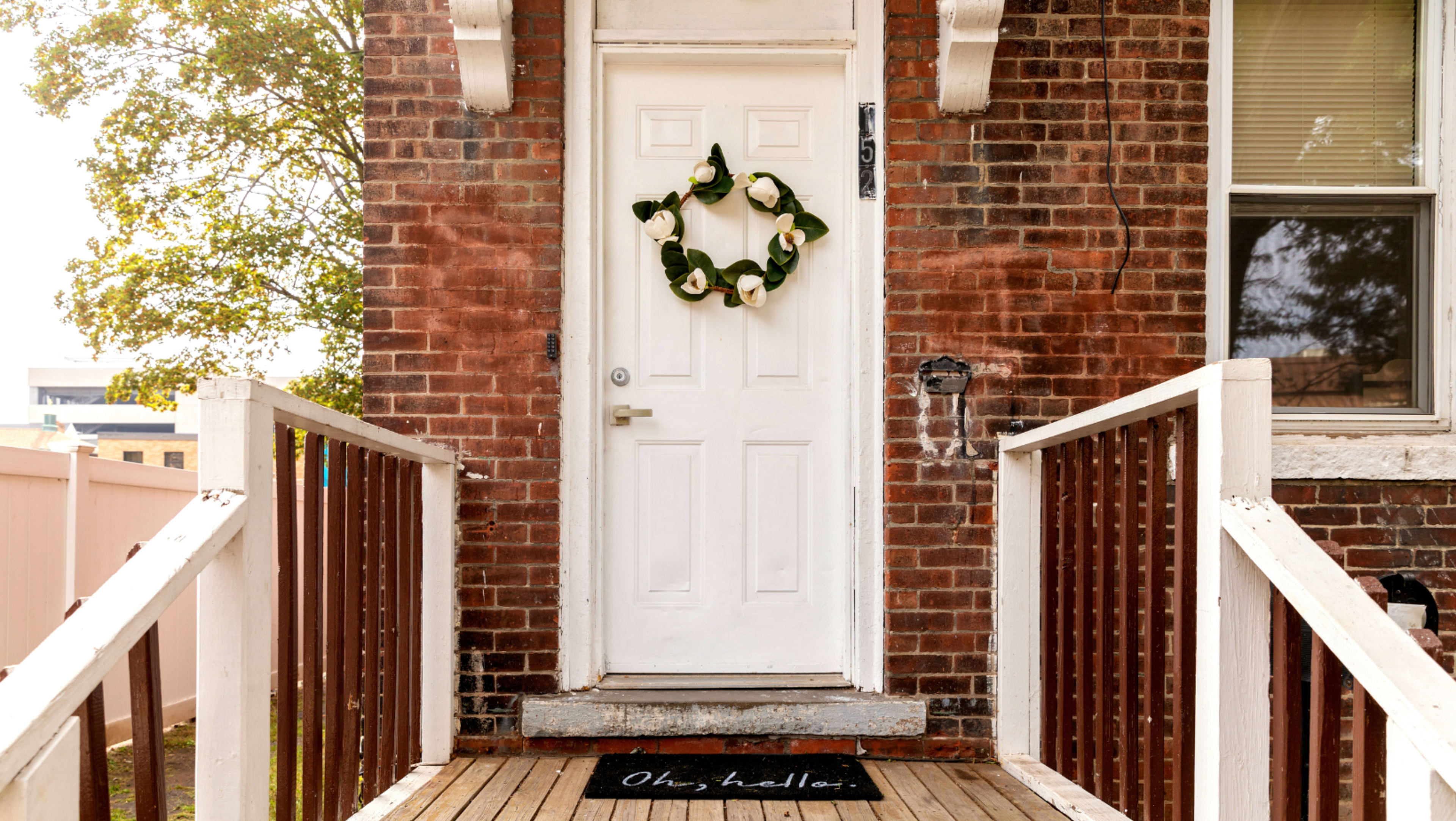 The image shows a white door adorned with a green wreath, set in a brick wall with a wooden steps leading up to it.