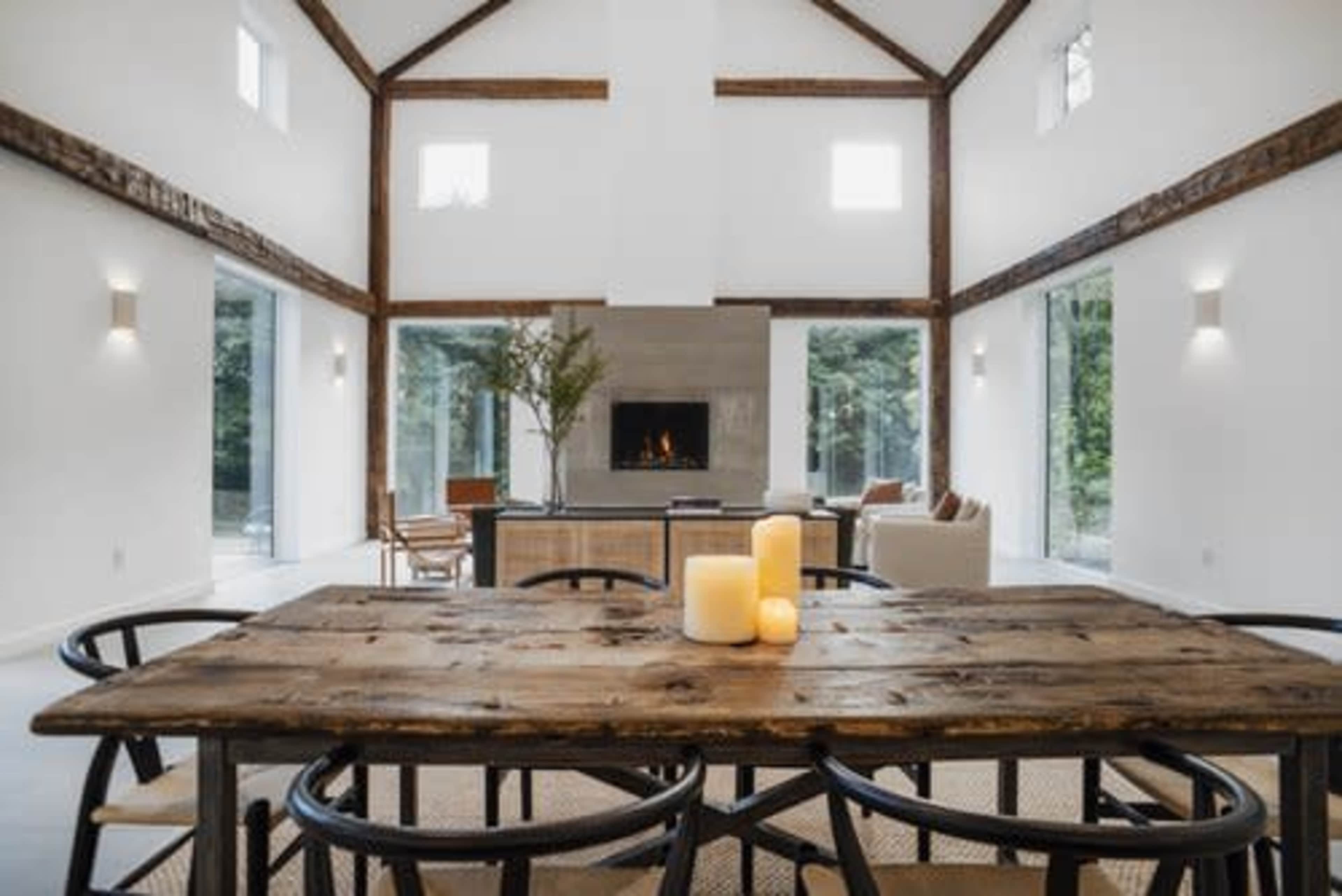 A spacious, modern living area features a wooden dining table with candles in the foreground and a fireplace surrounded by seating in the background.