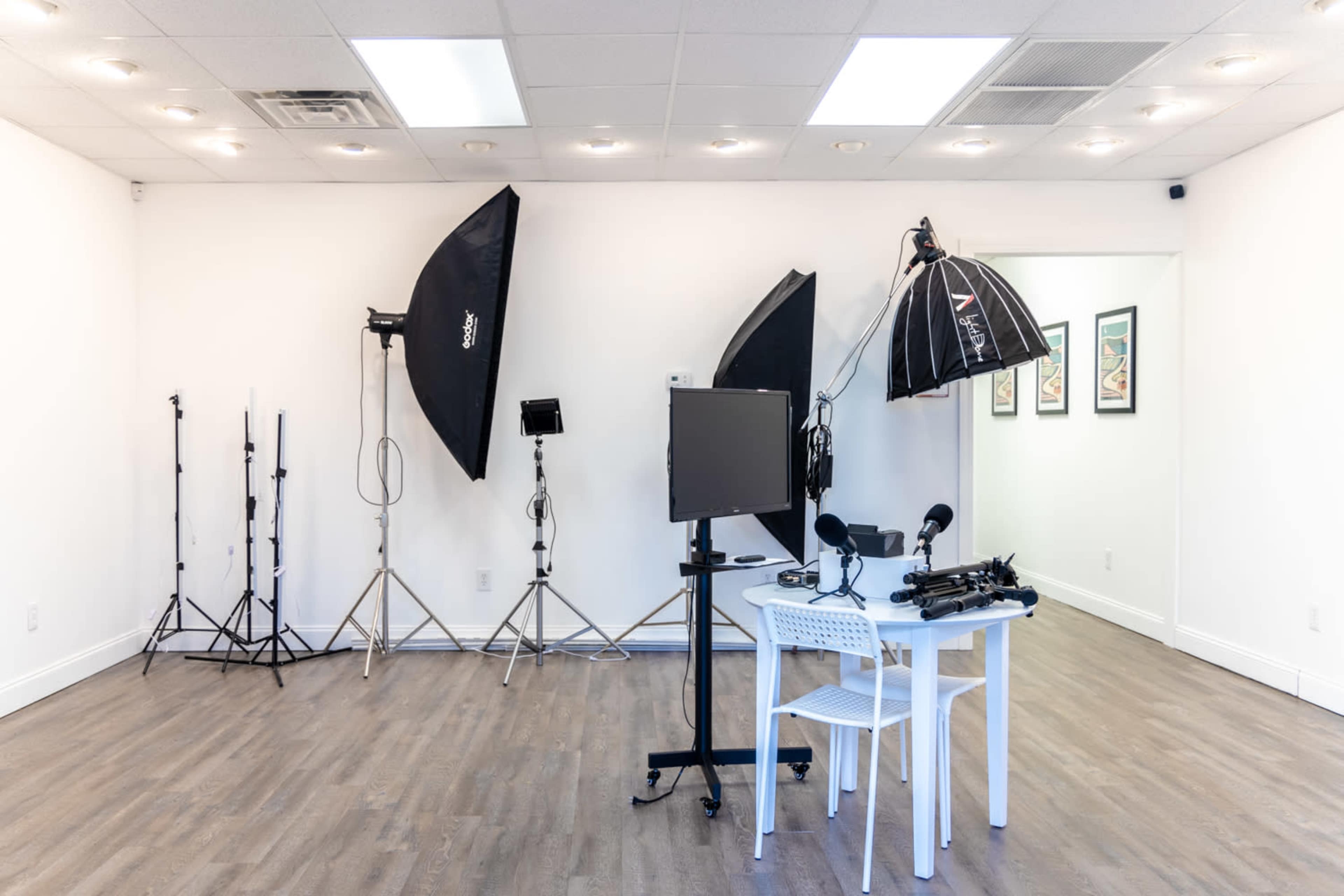 A bright, empty photography studio contains lighting equipment, a monitor, and a small table with microphones.