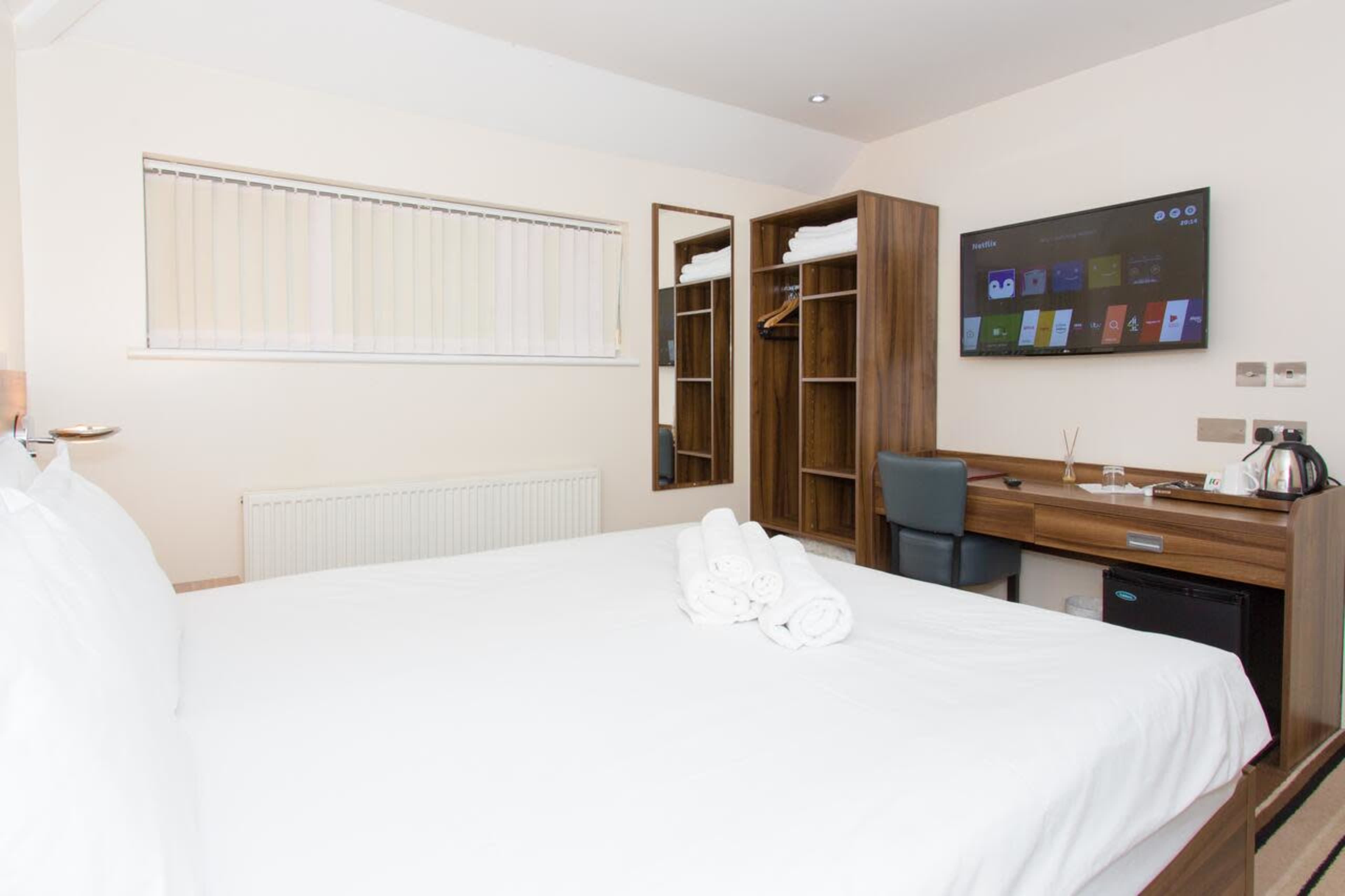 A neatly arranged hotel room features a double bed, a wooden wardrobe, a desk with a television, and a small refrigerator.