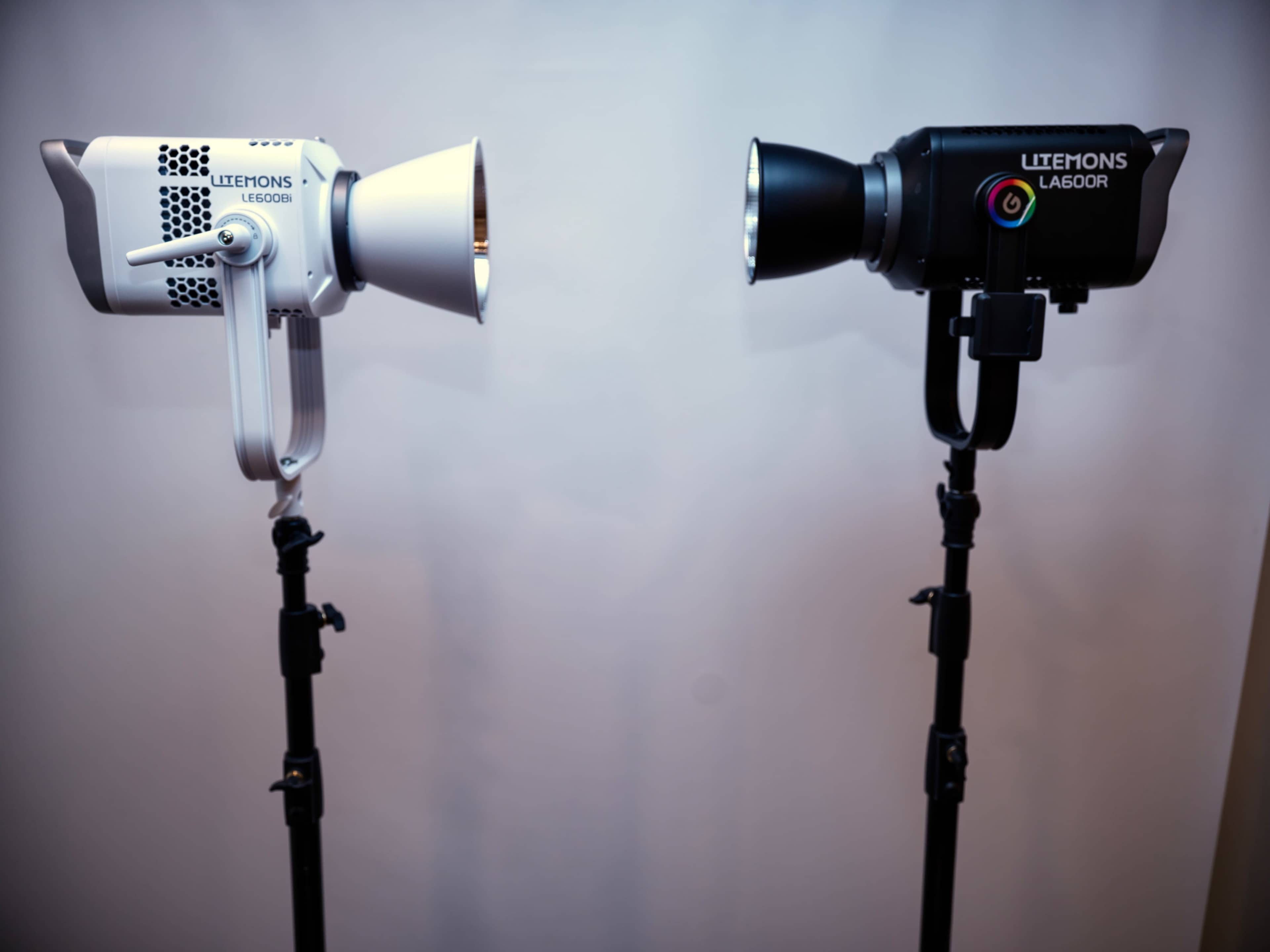 Two studio lights with different designs are mounted on adjustable tripods against a plain background.