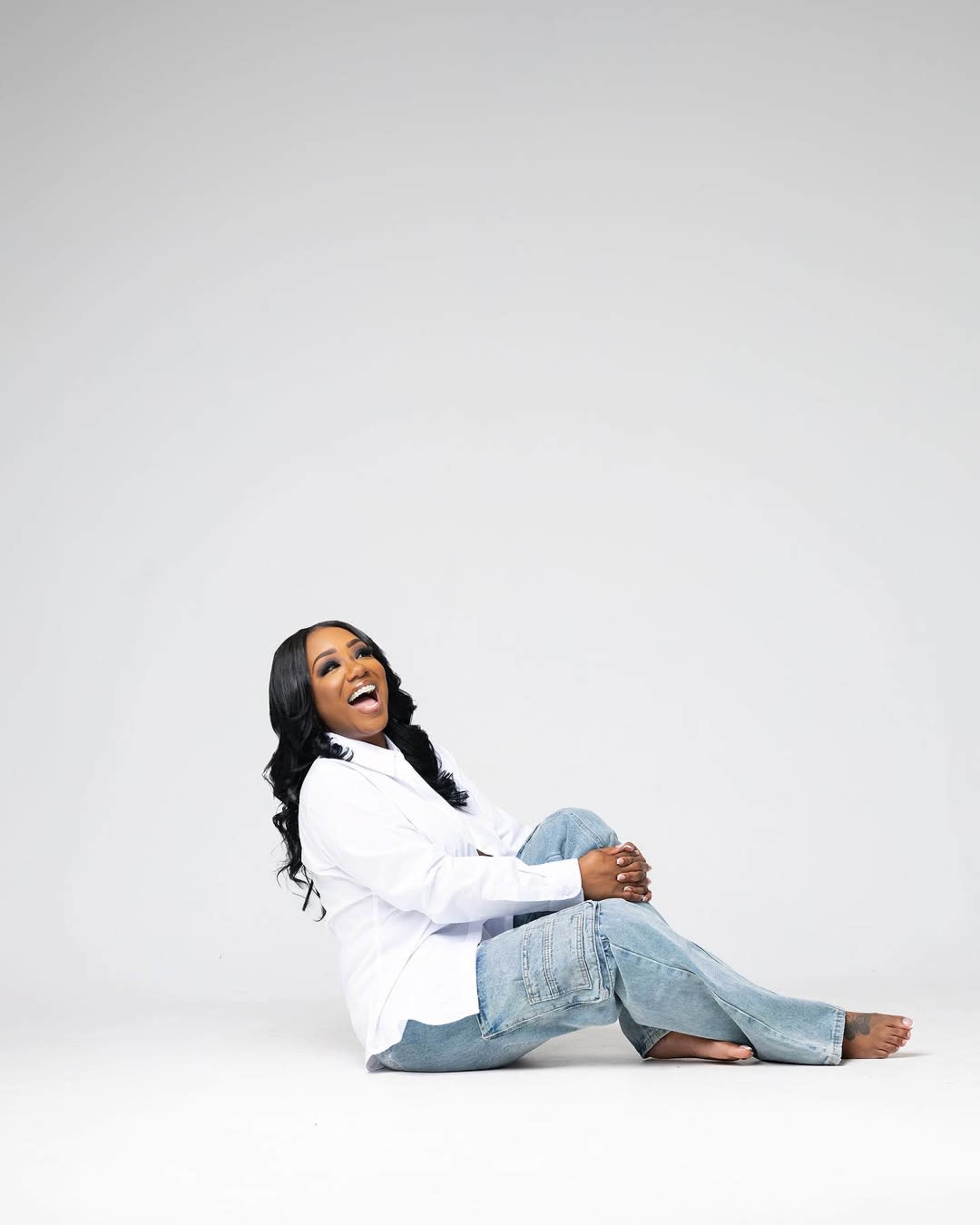 A woman sits on the floor, smiling while wearing a white shirt and denim jeans against a plain white background.
